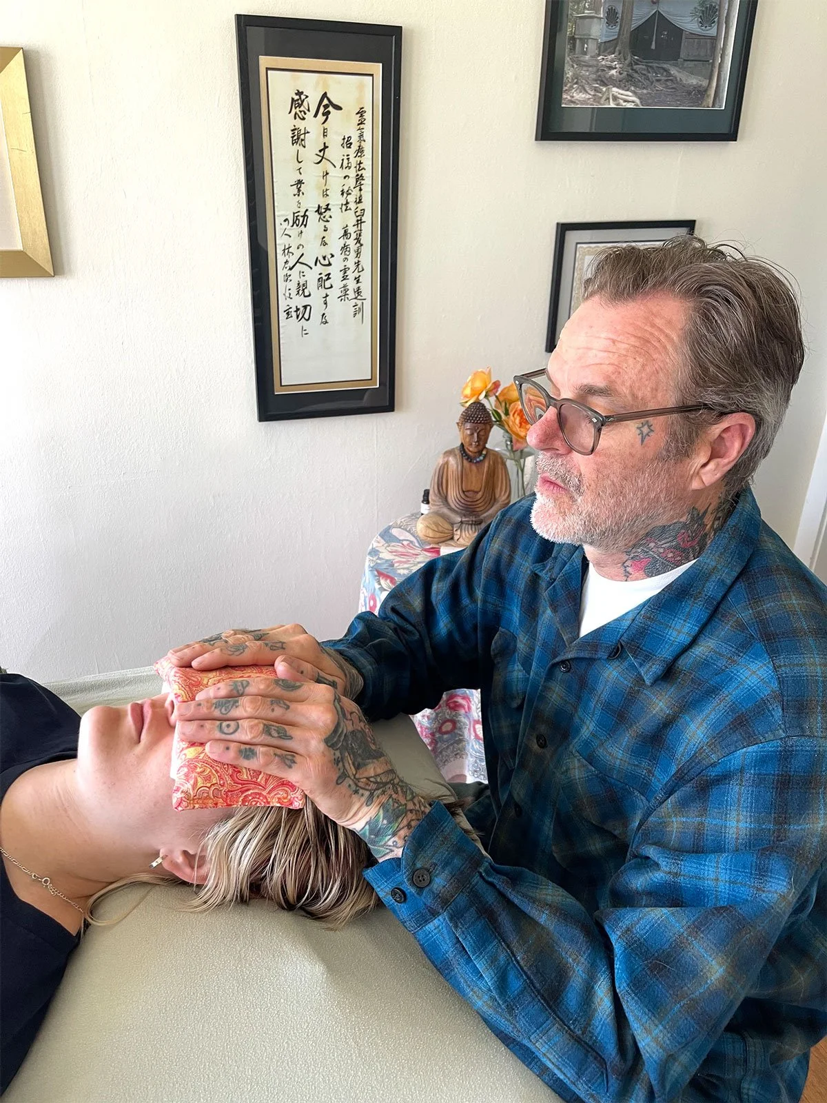 A man receiving a facial treatment from another man with tattoos on his hands and neck, in a room decorated with Asian art, a Buddha statue, and a small table with flowers.