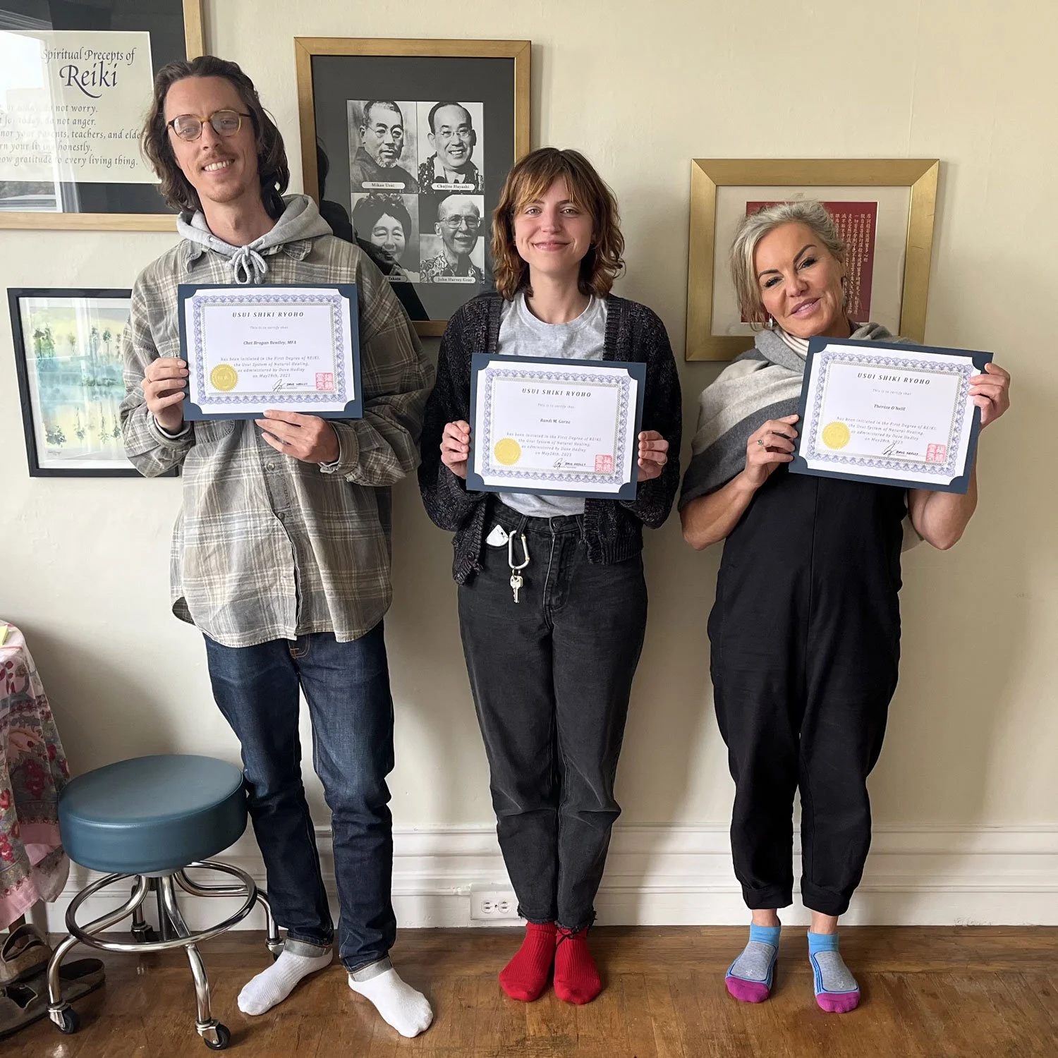 Three people standing side by side holding certificates, smiling for the photo. The person on the left is wearing glasses, a plaid shirt, and dark jeans. The person in the middle has shoulder-length hair, wearing a black cardigan, a gray t-shirt, and