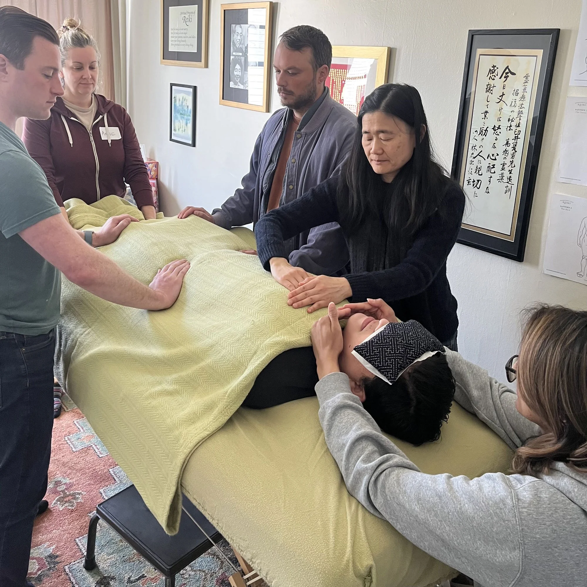 People gathered around a massage table, with one person receiving a massage from a woman practicing energy healing techniques in a room decorated with framed artwork.