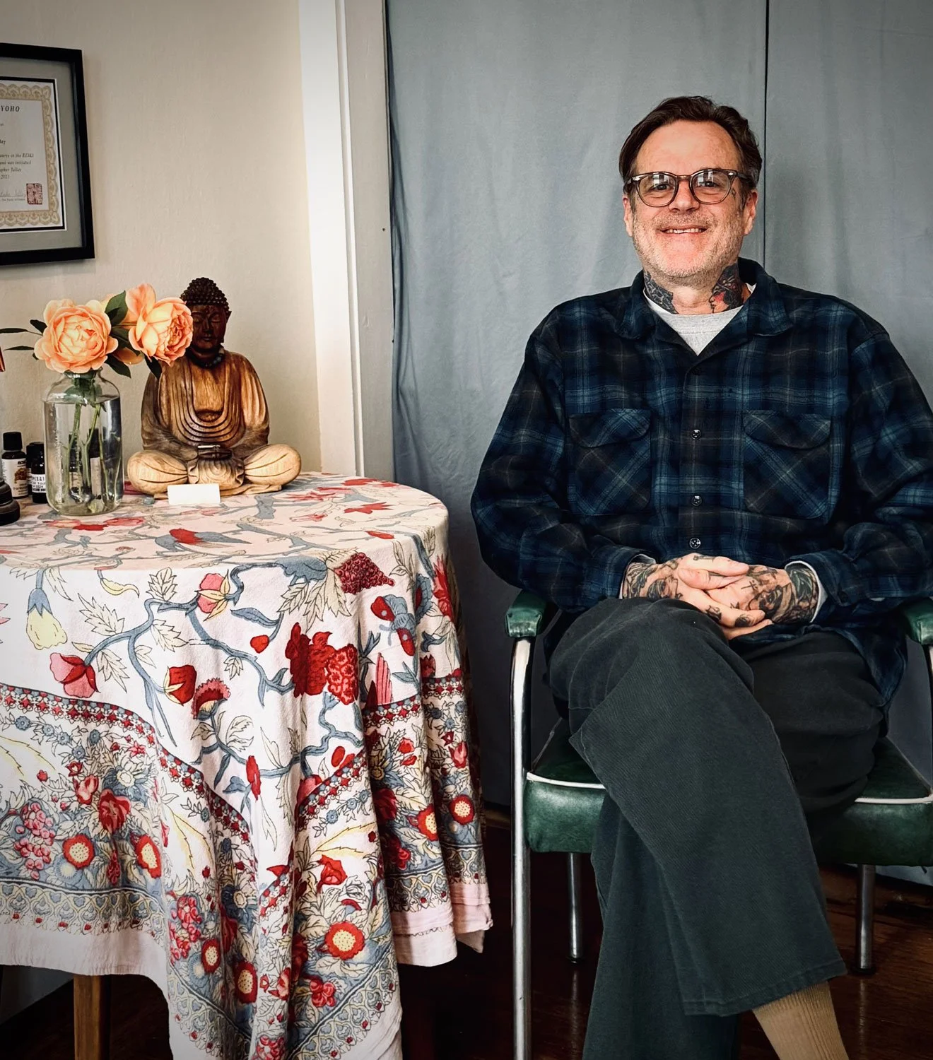 A man with glasses and tattoos sitting in a chair, smiling, wearing a black and blue plaid shirt and black pants, with a table beside him decorated with a floral tablecloth, a Buddha statue, a vase with pink roses, and various small bottles.