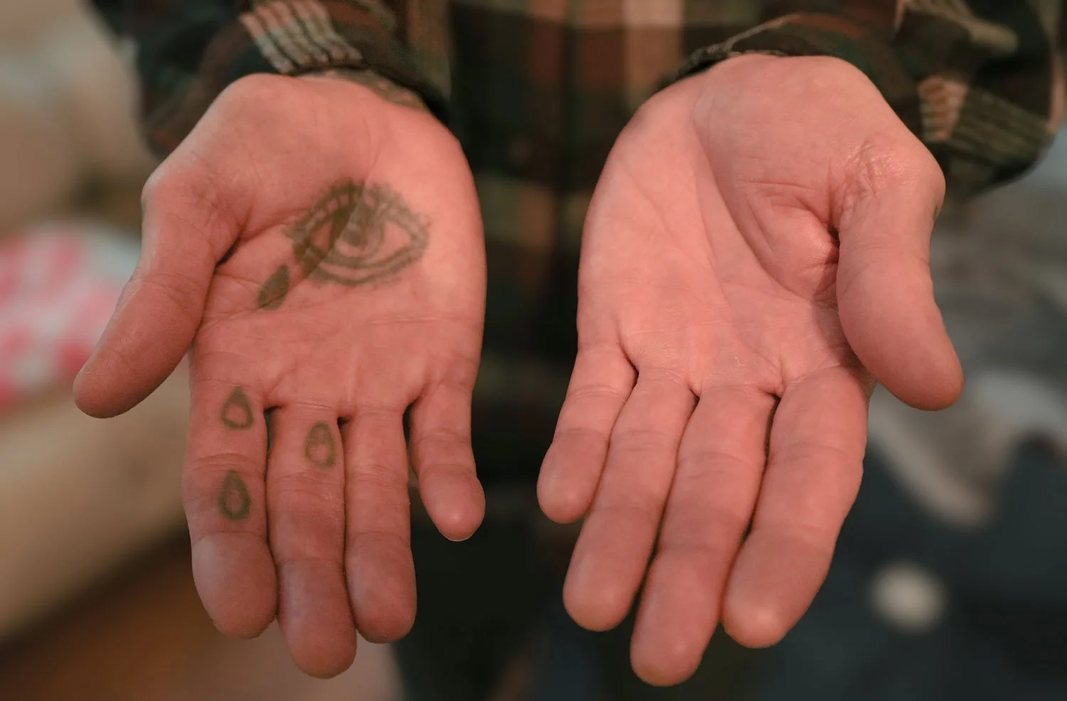 Person showing their open hands, one with a tattoo of an eye and teardrops, the other hand plain.