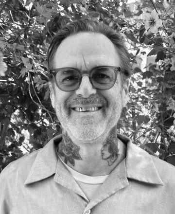 Black and white photo of a smiling man with glasses, tattoos, and a collared shirt standing outdoors with leafy foliage in the background.