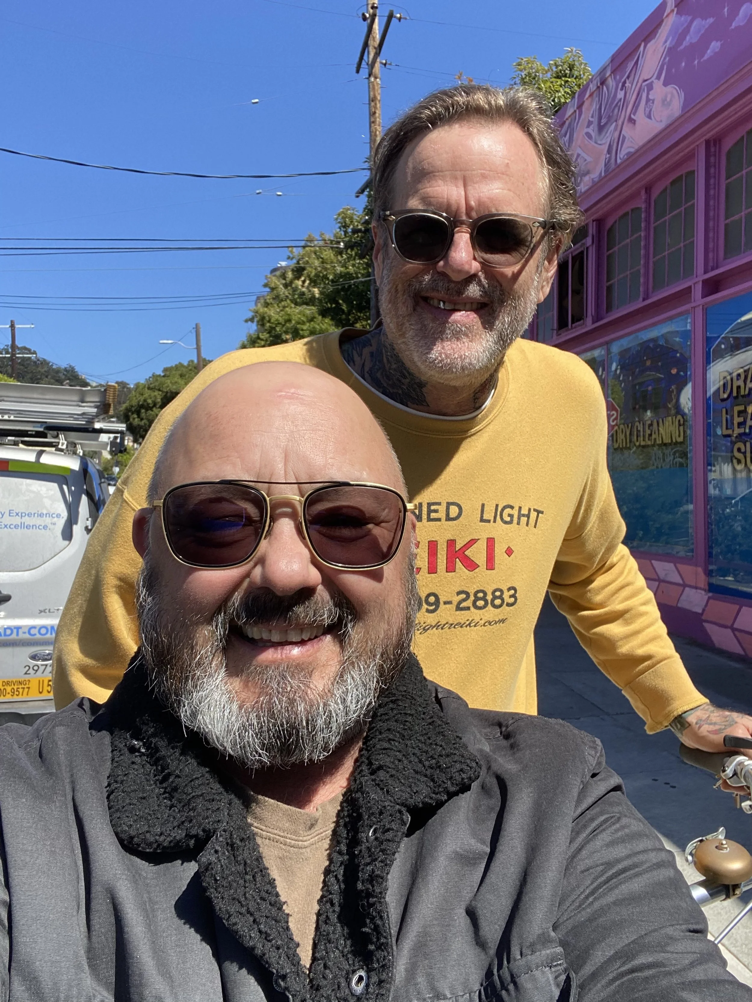 Two smiling men wearing sunglasses taking a selfie on a sunny day, with a pink storefront and a utility pole in the background.