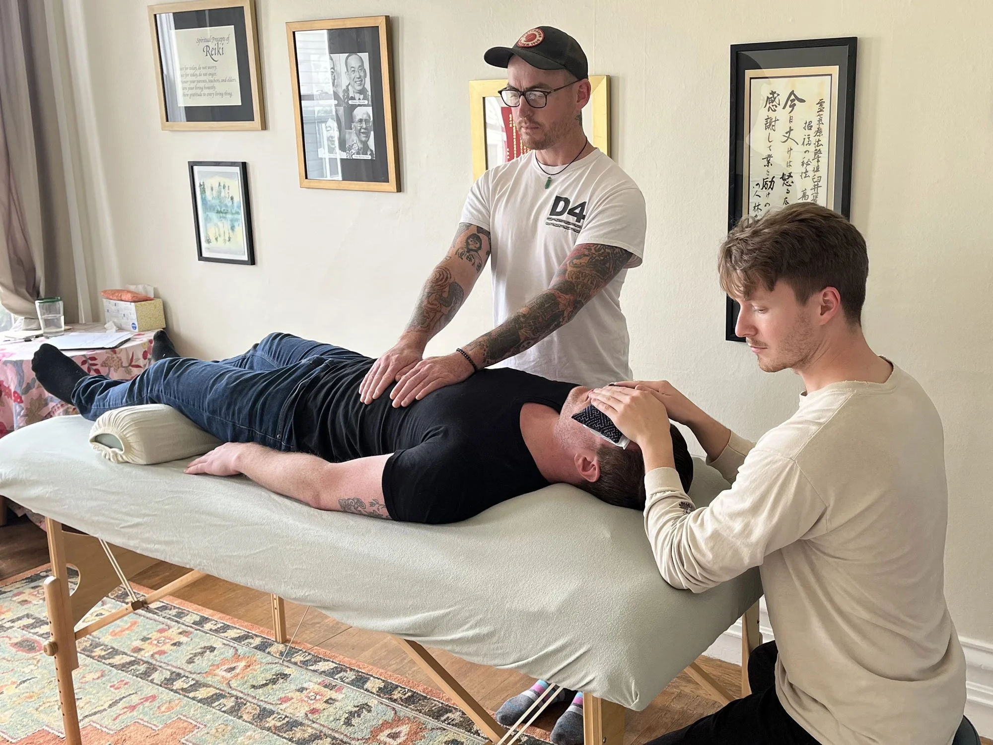 Two men giving a massage to a third man lying face down on a massage table, with a third person checking his forehead. The scene takes place in a room with framed artwork on the wall.