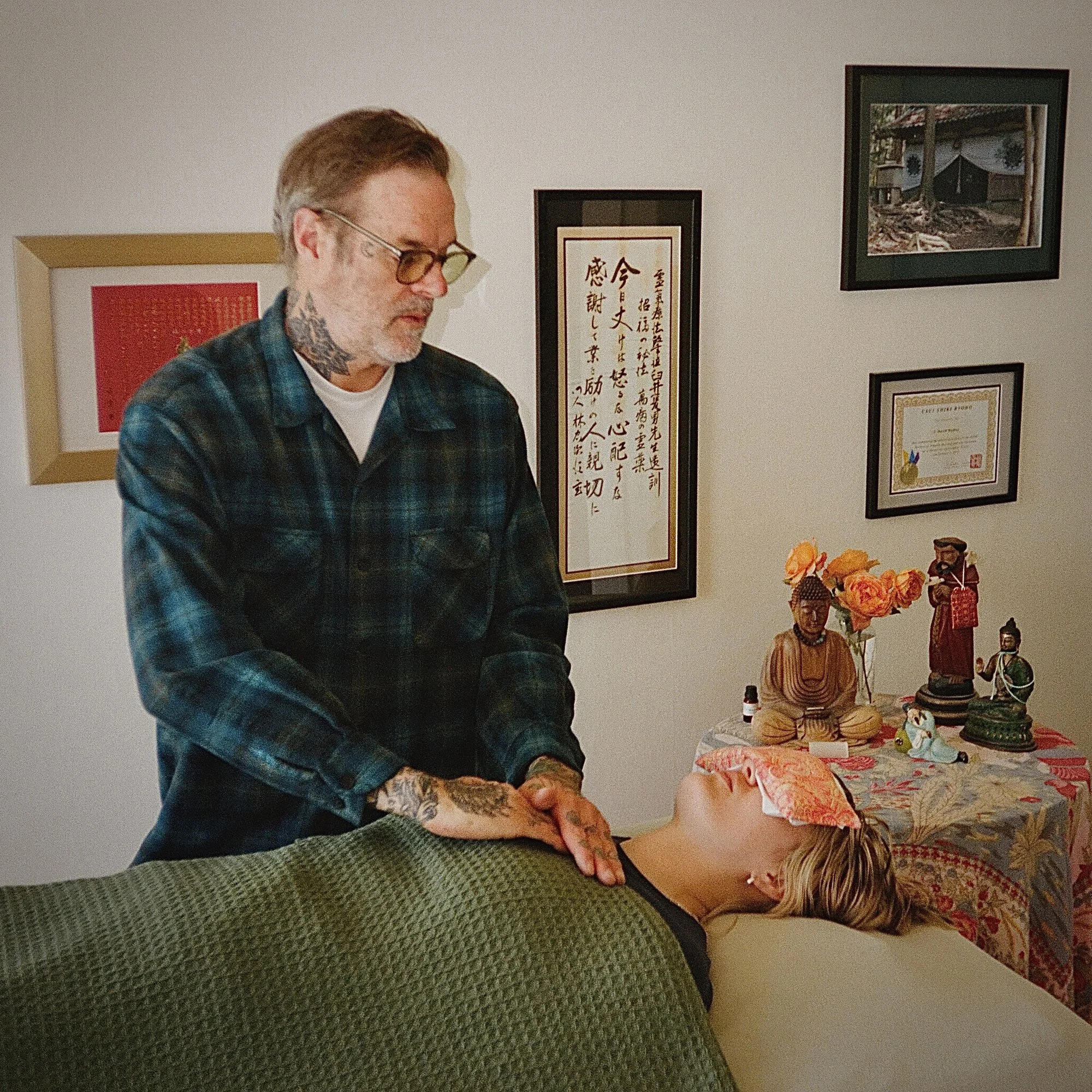 A man with glasses, tattoos, and a plaid shirt performs Reiki on a woman lying on a massage table, who has a pink cloth over her eyes.