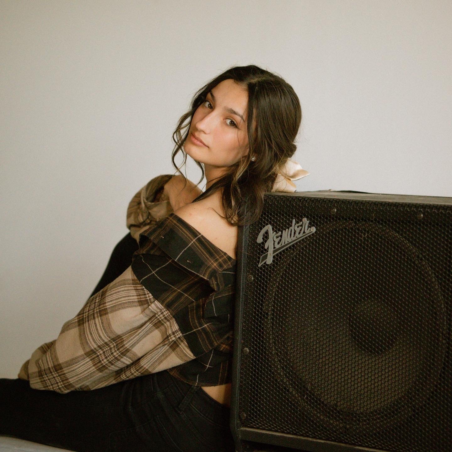 A young woman with shoulder-length wavy dark hair, wearing a plaid shirt, leaning against a large Fender speaker, with a plain light-colored wall in the background.