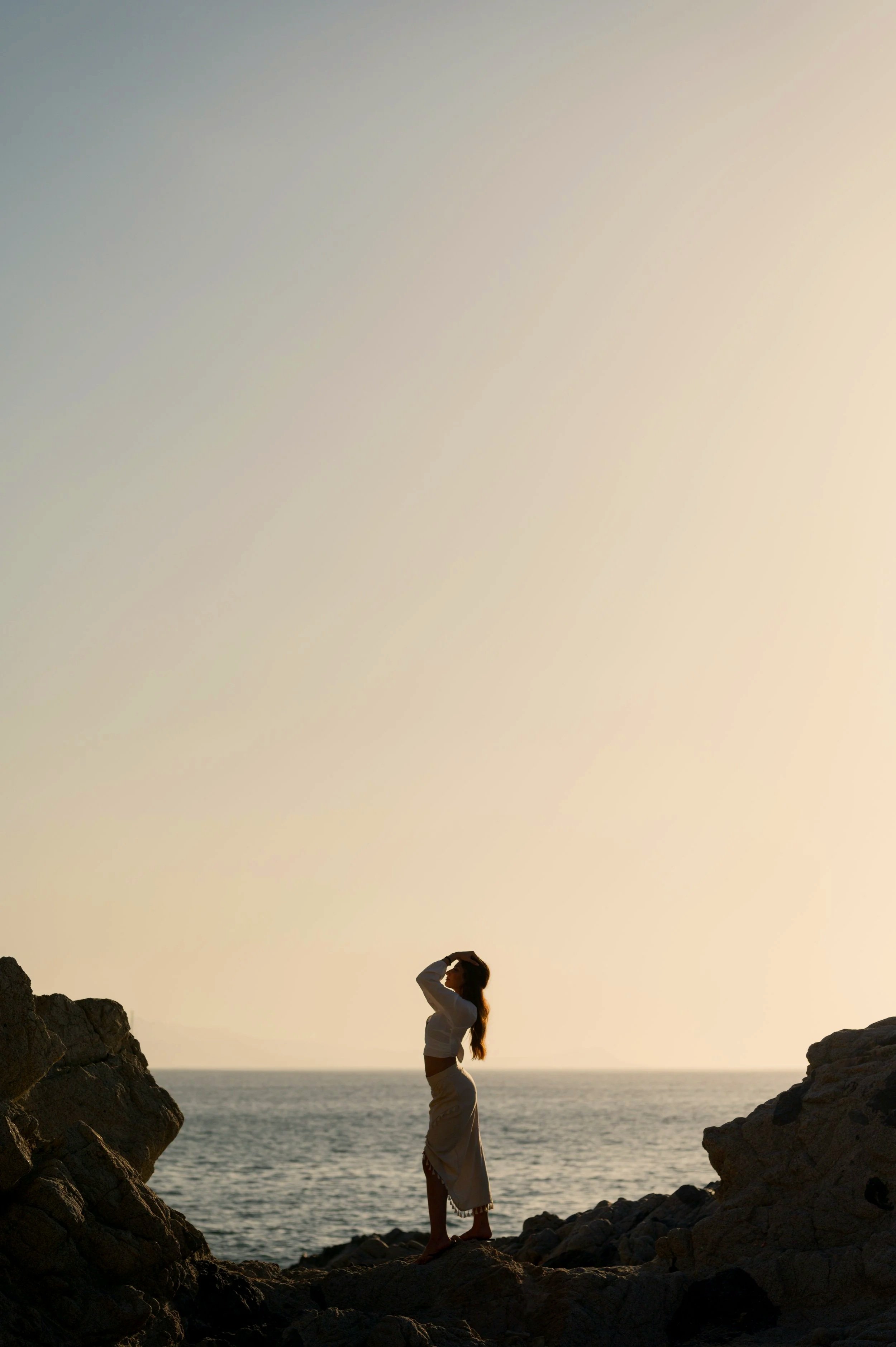 Silhouette of a woman standing on rocks at the beach with the ocean in the background during sunset or sunrise.