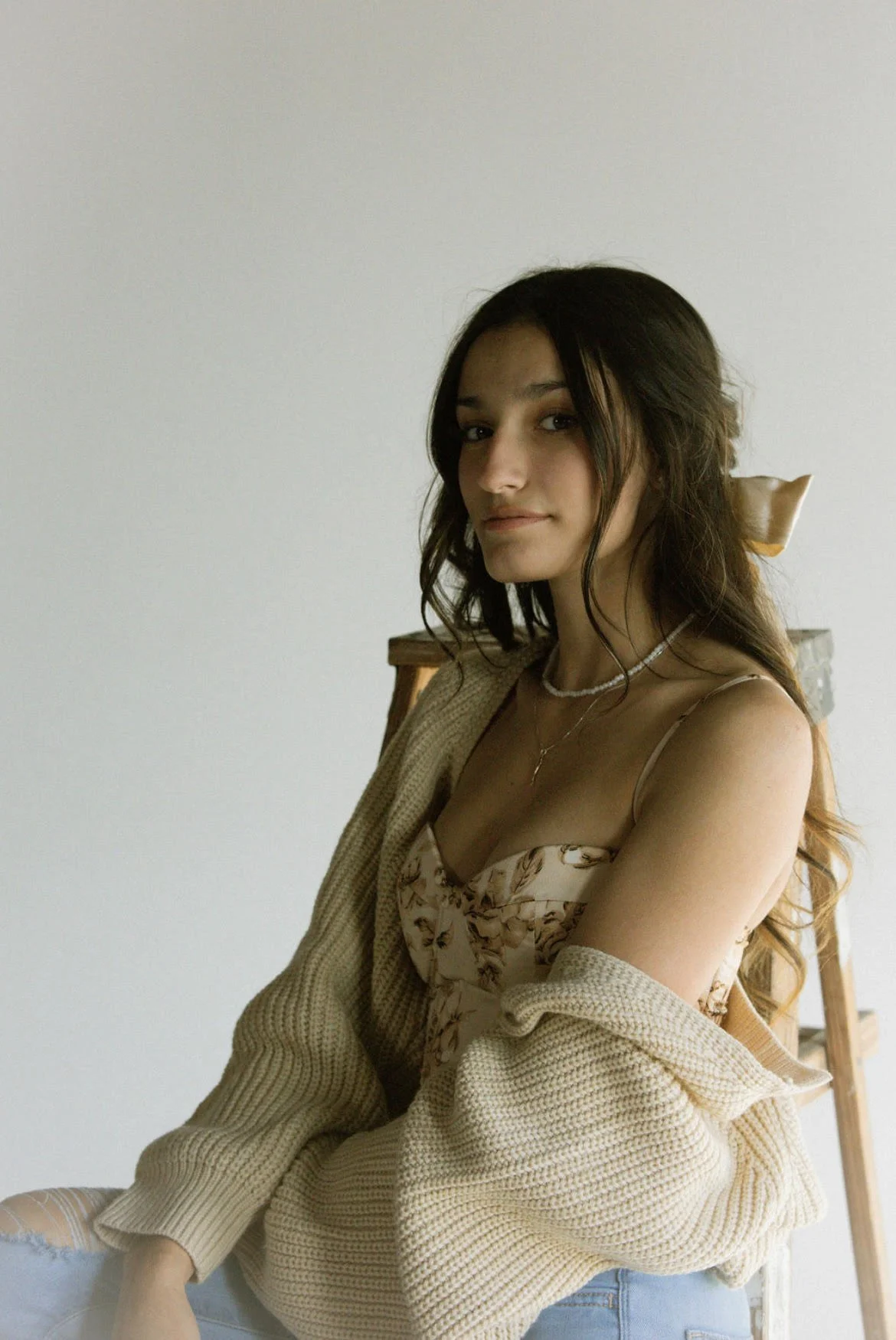 A young woman with long brown hair sitting on a chair, wearing a beige cardigan, floral slip dress, layered necklaces, and ripped jeans, against a plain white wall.