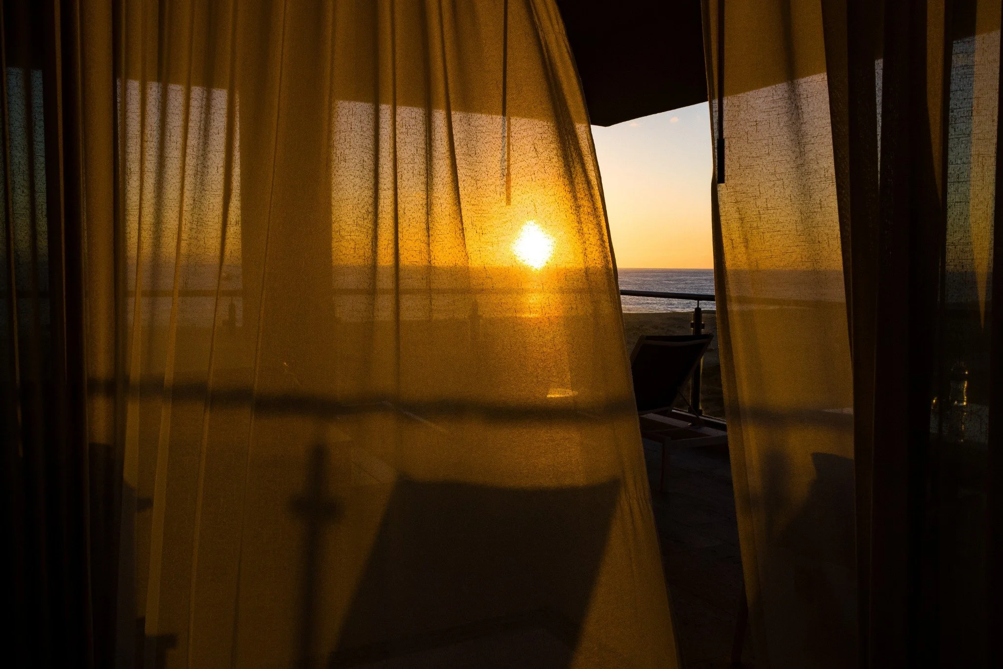 Sunset view through yellow curtains on a balcony near the beach with a lounge chair.