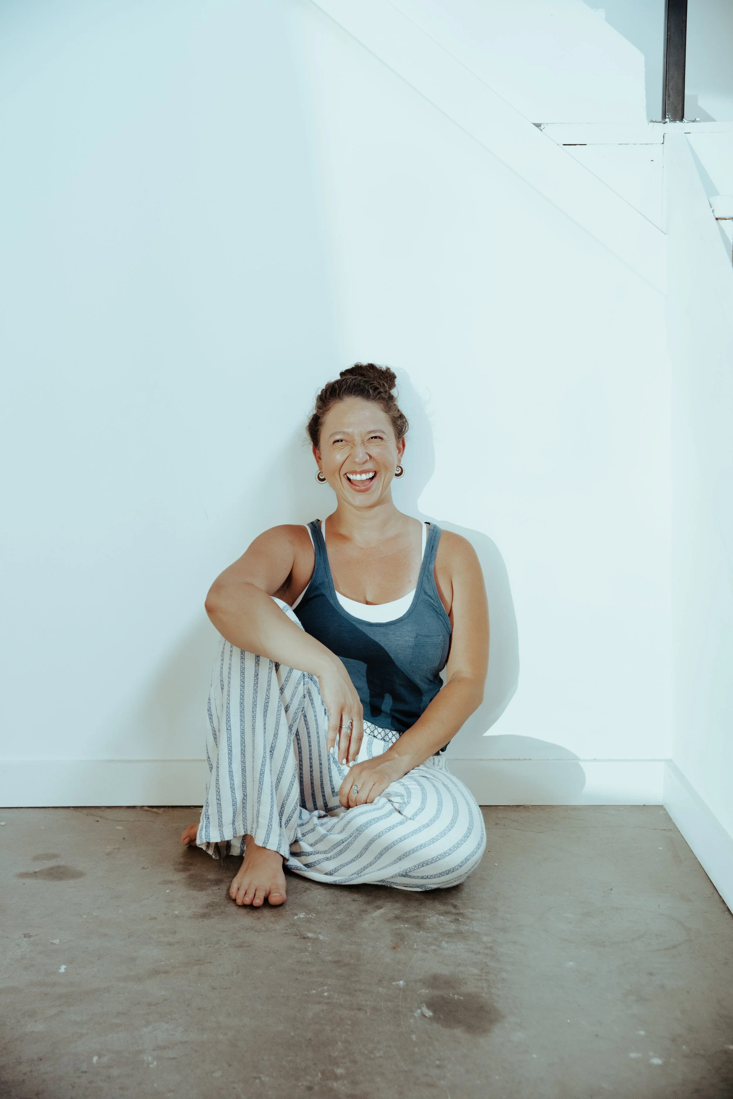 Woman sitting on the floor against a white wall, smiling and laughing, dressed in pajamas.