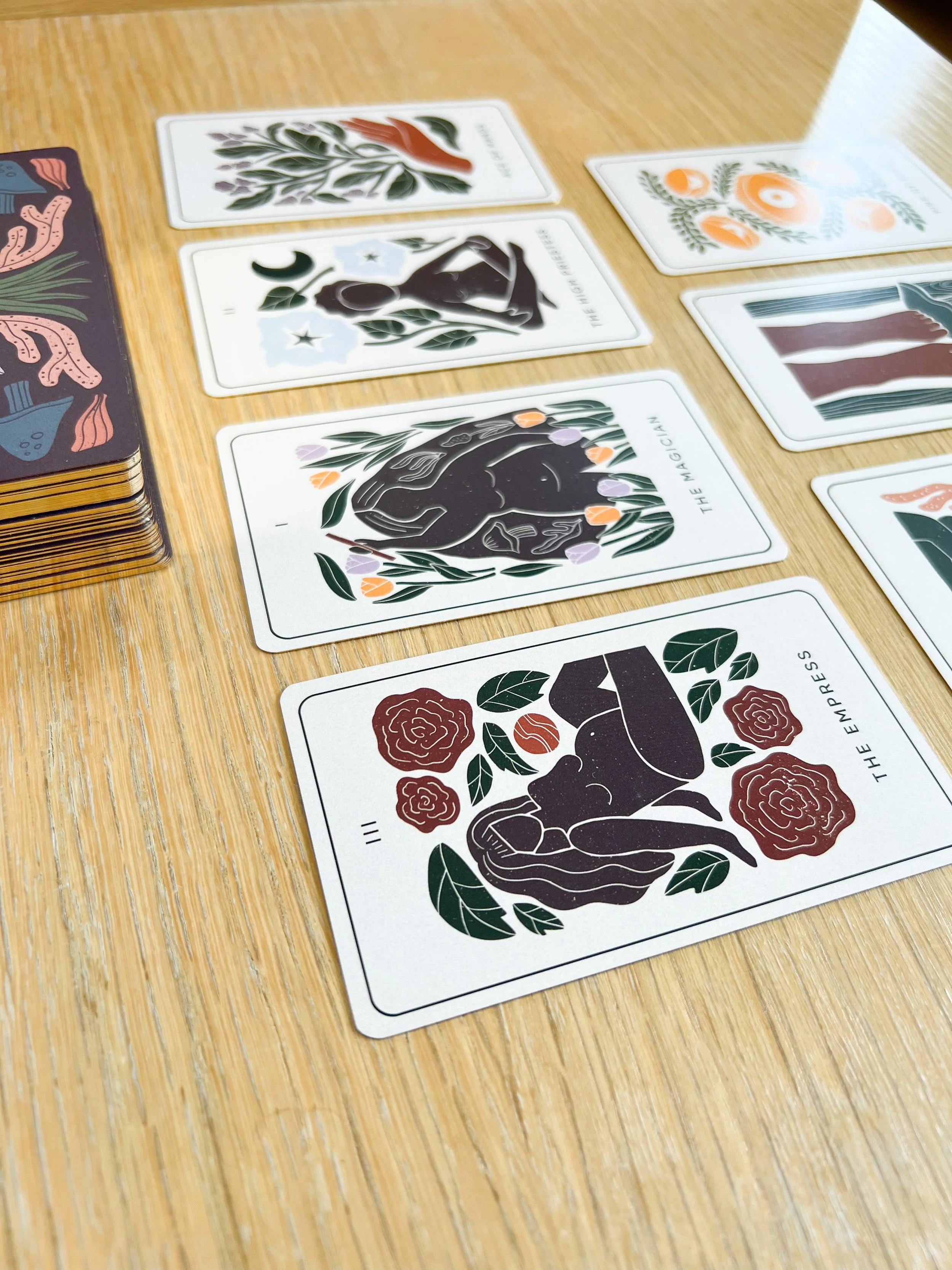 A tarot card game laid out on a wooden table, featuring cards with stylized illustrations of figures, symbols, and flowers in black, white, and earthy colors.
