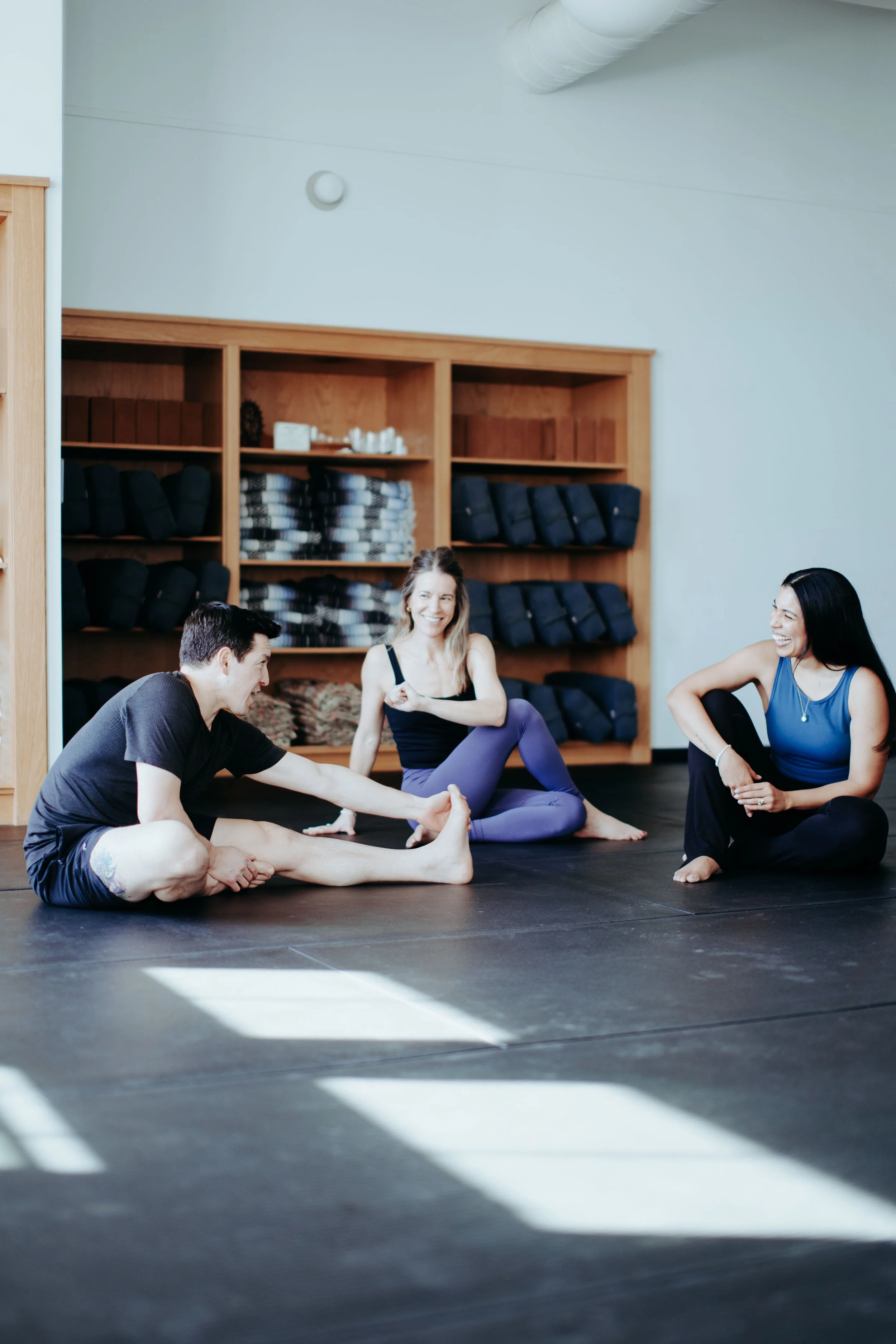Three people sitting on yoga mats in a studio, laughing and stretching during a yoga class.
