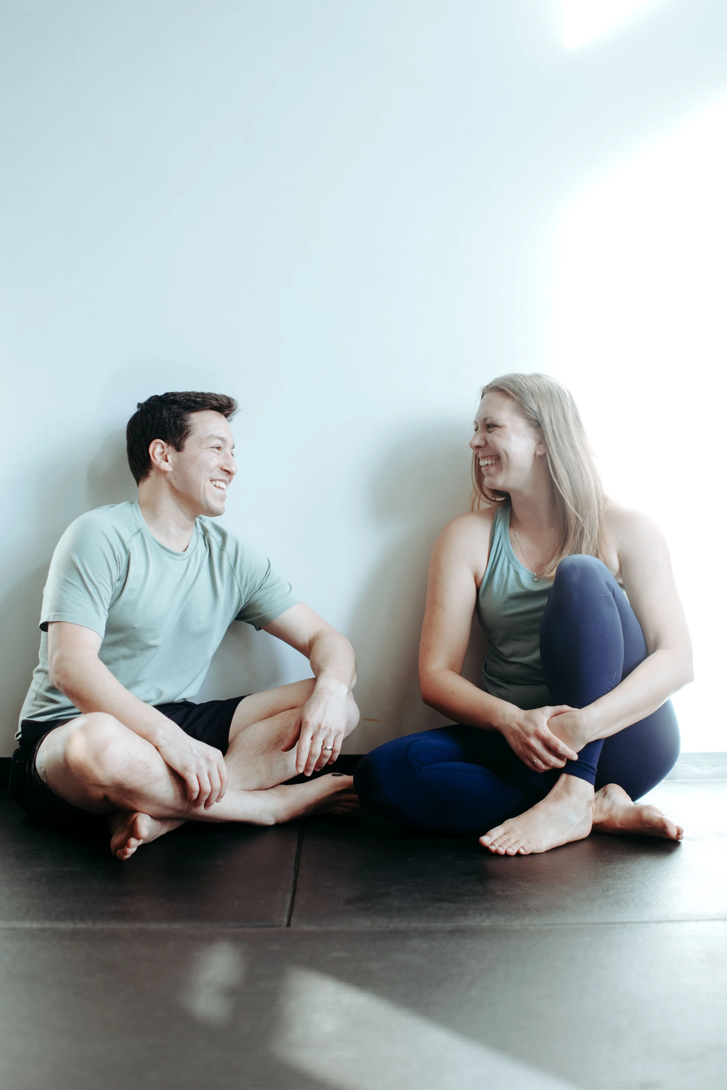 Two people sitting on a black mat inside, smiling and talking, one man and one woman in casual workout clothes, near a plain white wall, enjoying a relaxed moment.