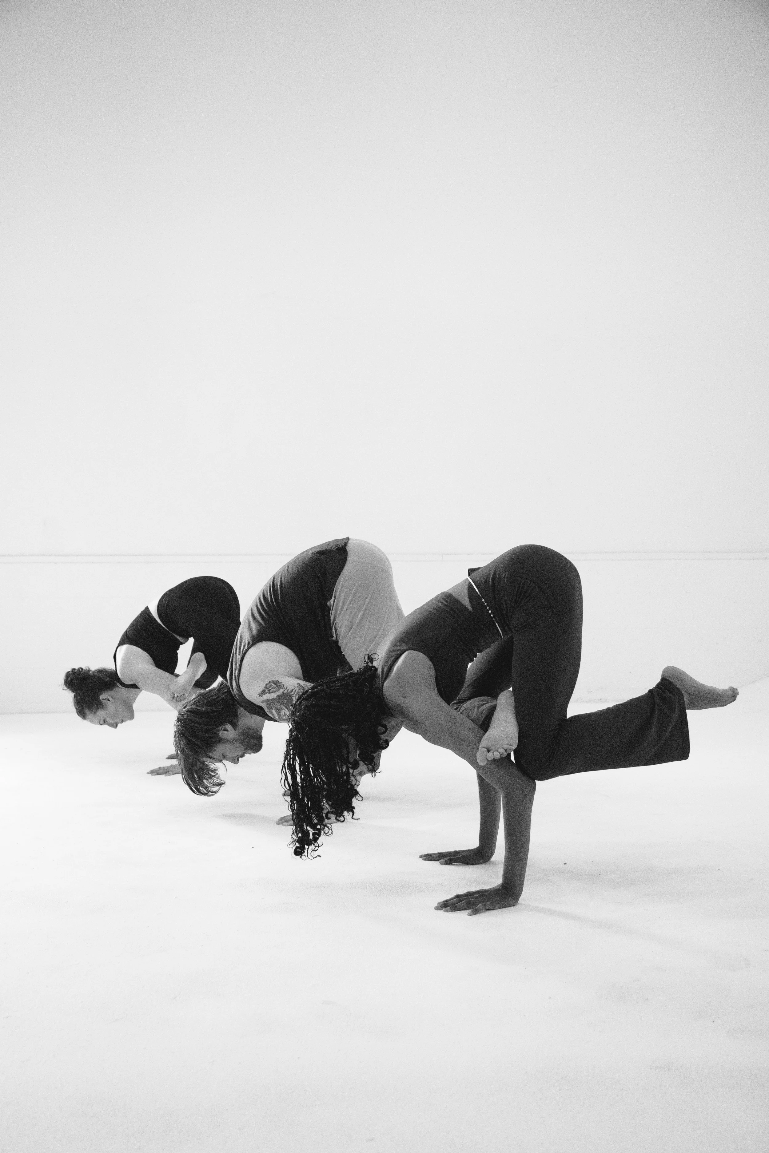 Four women practicing acro yoga or partner yoga in a gym or studio, performing a complex balancing pose with two in a plank position and two balancing on their shoulders and legs.