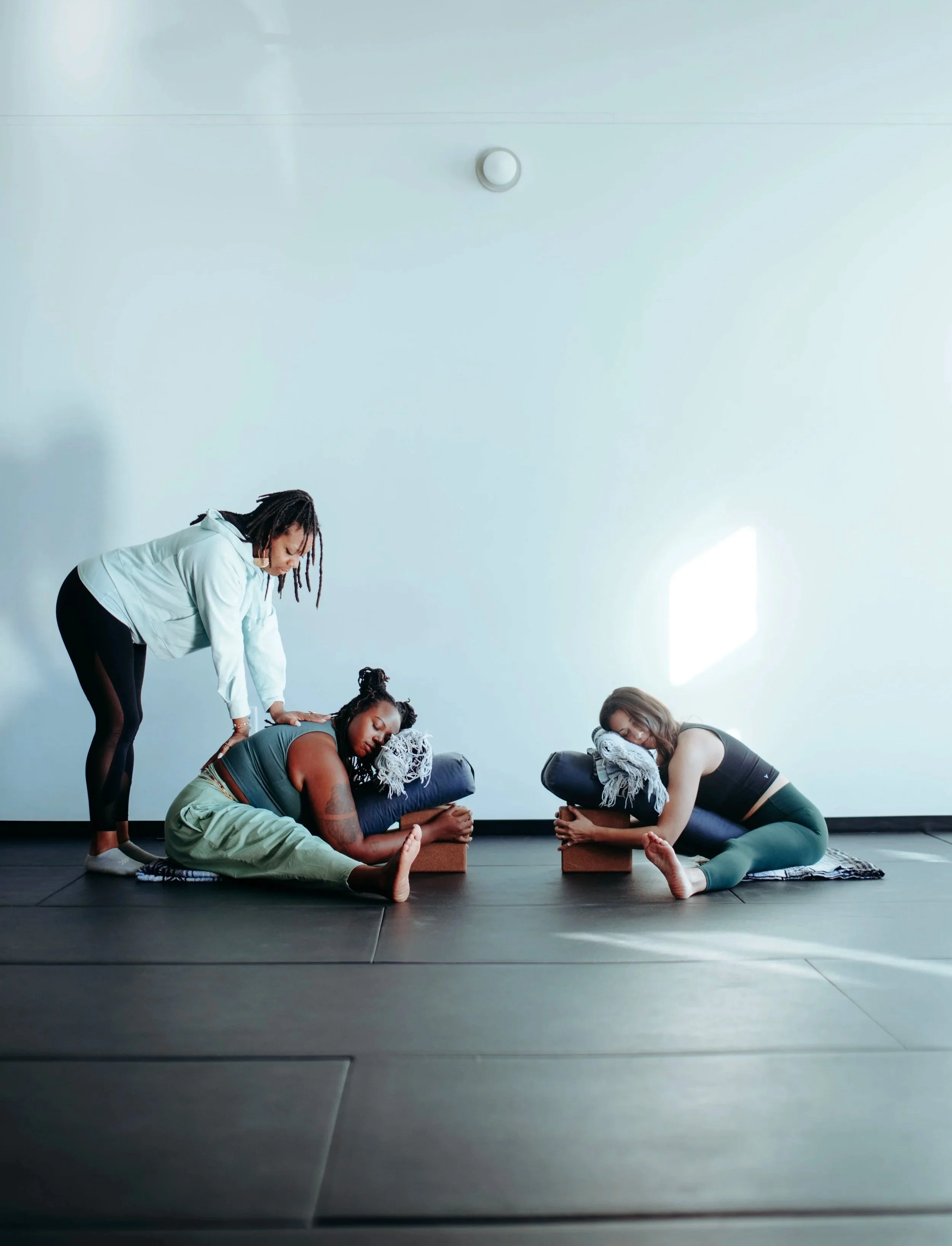 Two women practicing yoga with bolsters and blankets in a bright, minimal studio, assisted by a instructor.