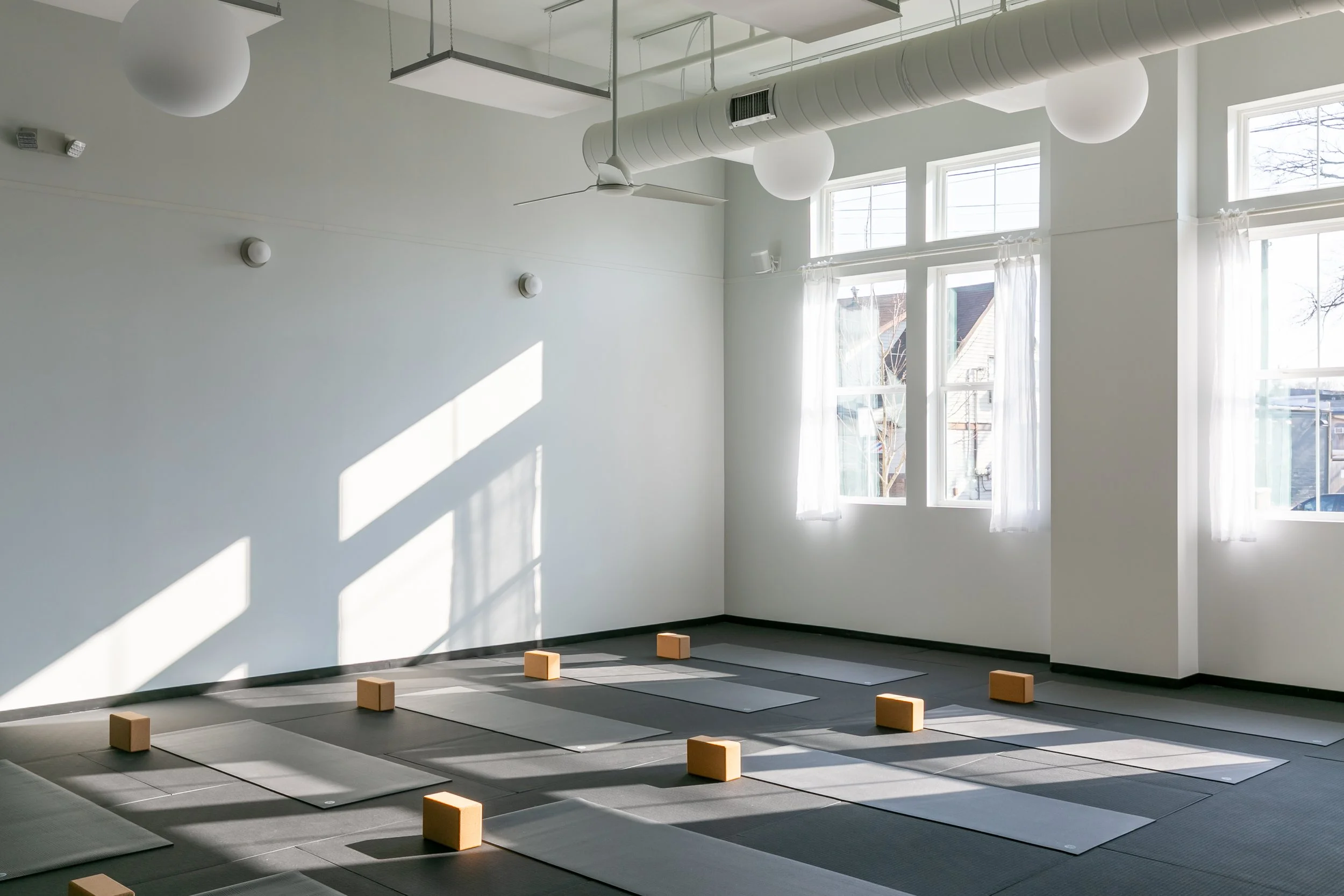 An empty yoga studio with sunlight streaming through large windows, yoga mats arranged on the floor with small wooden blocks, a ceiling fan, and white walls.