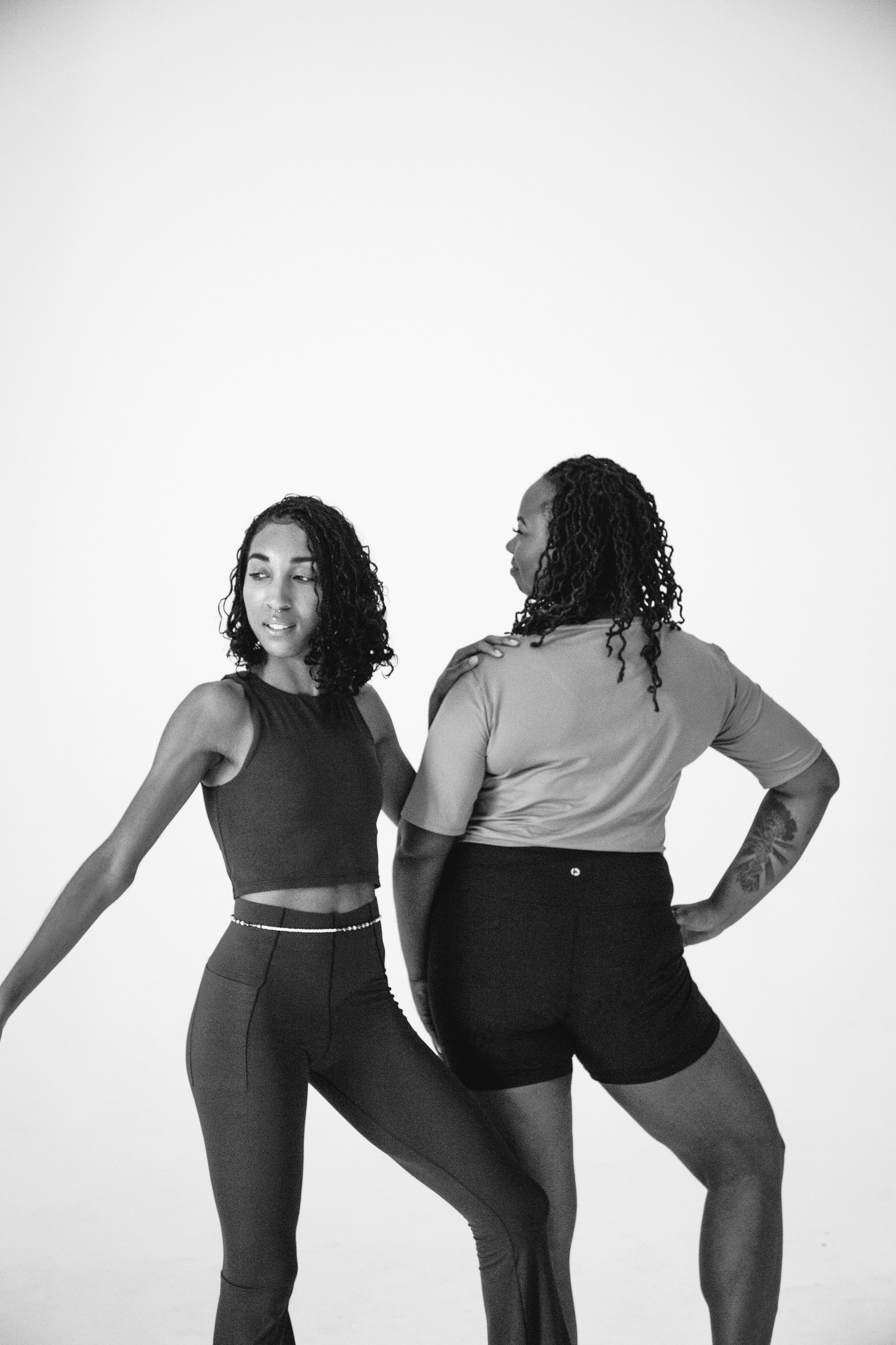 Two women standing together, one with her hand on her hip and the other with her arm extended, in a black-and-white photo against a plain light background.