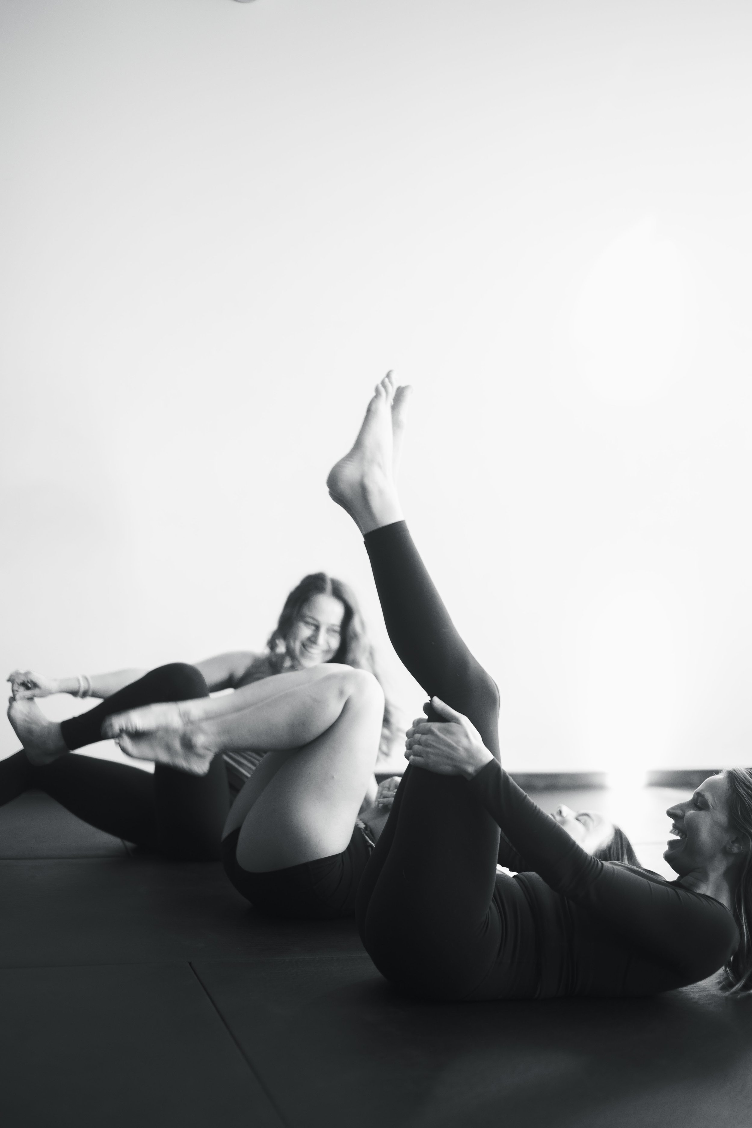 Two women in black workout clothes participate in a laughing yoga or stretching session on a mat, one lying on her back and the other sitting on her feet. The scene is in a bright, minimal studio.