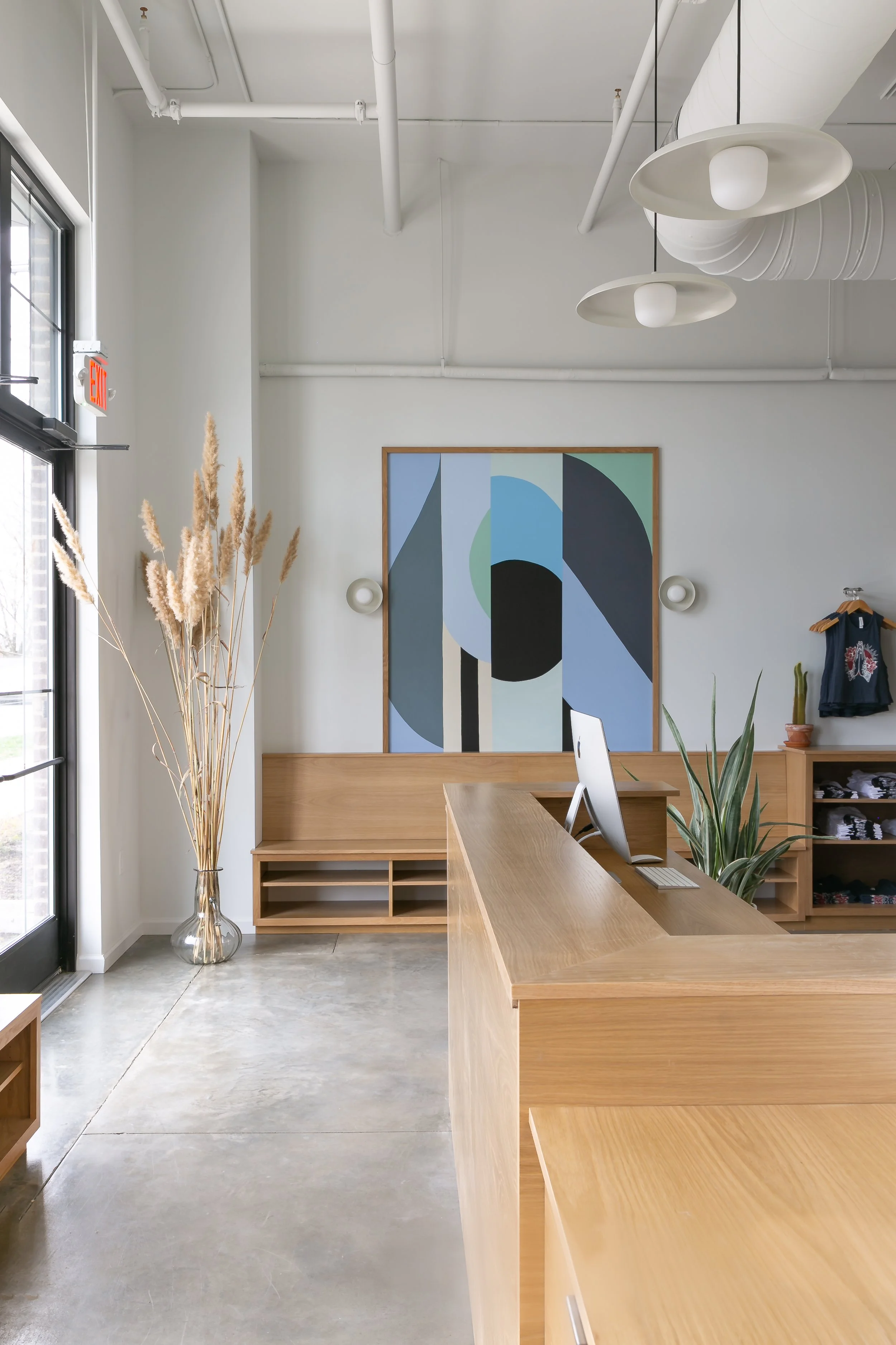 Modern reception area with a wooden counter, a computer, large abstract artwork on the wall, tall dried pampas grass in a glass vase, a potted plant, and clothing on a small rack, illuminated by natural light from glass doors.