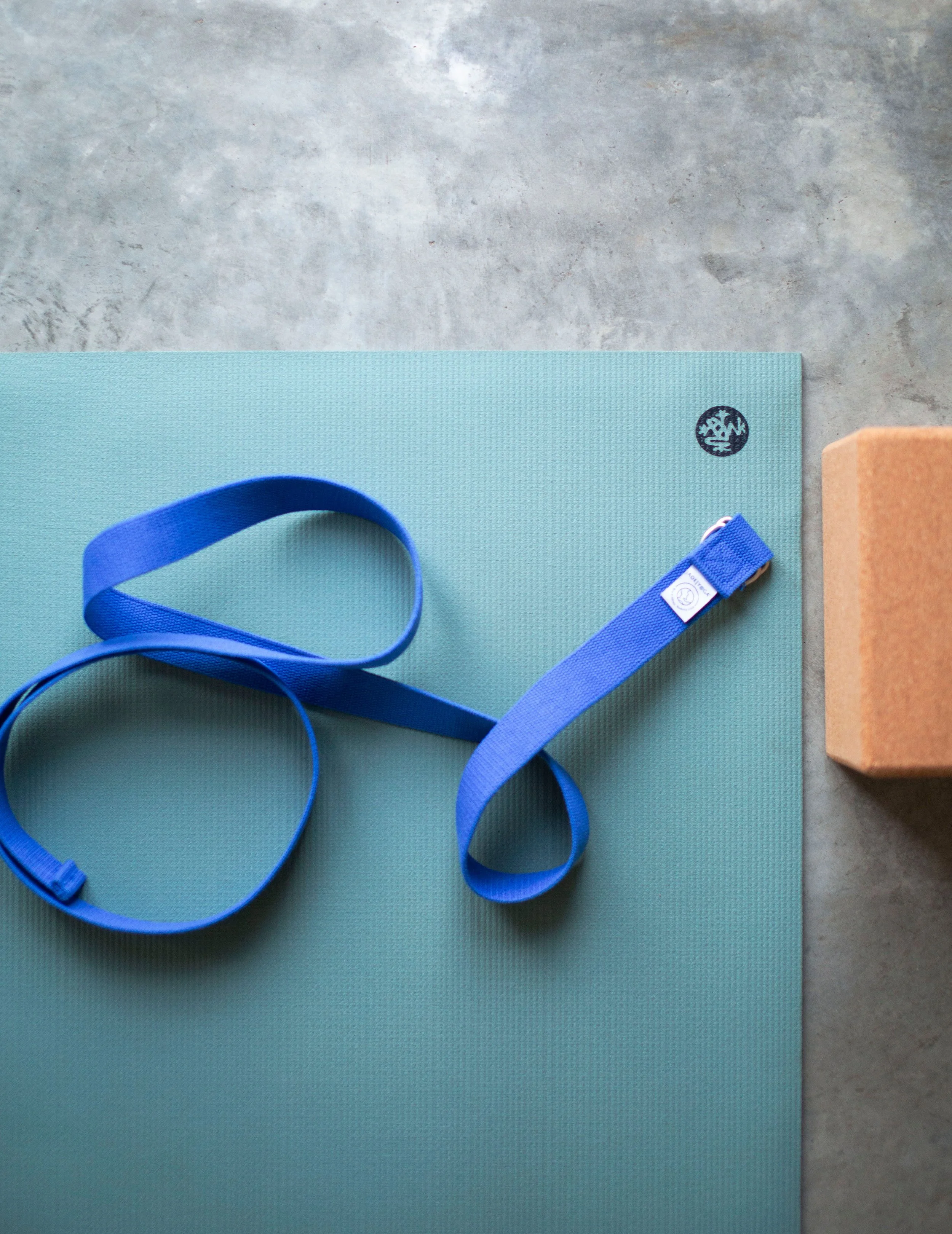 A blue yoga strap on a light blue textured yoga mat with a small black logo, next to a cork yoga block on a gray surface.