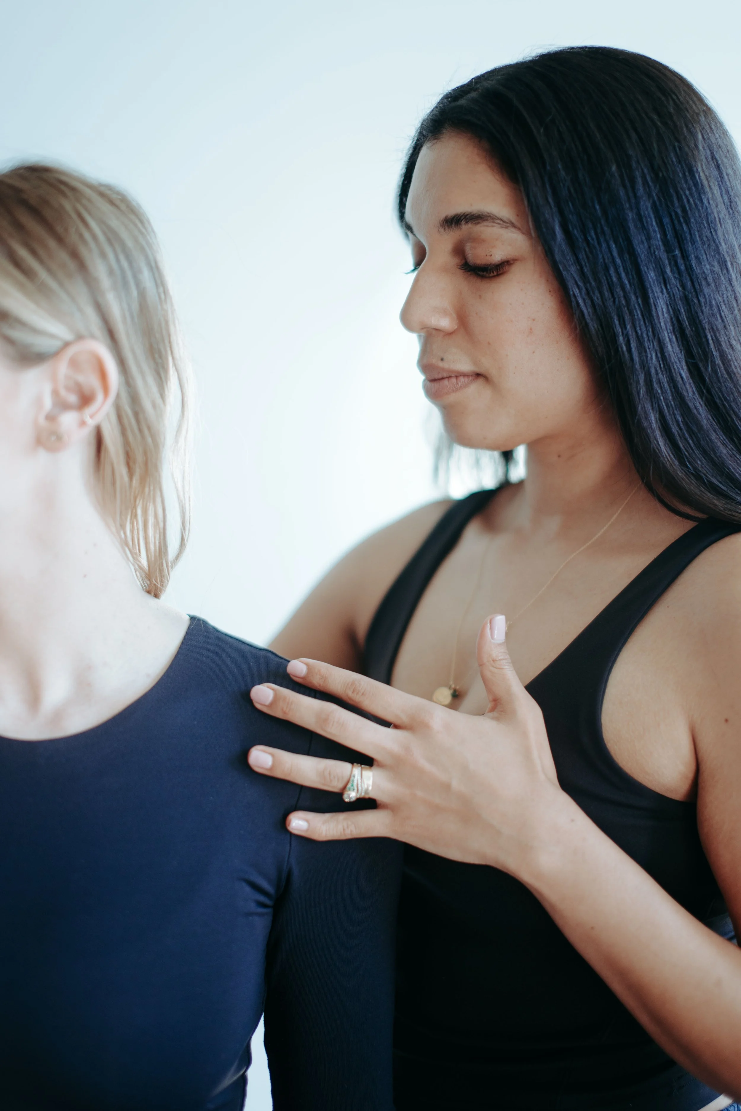 Two women are engaging in a spiritual or emotional healing session, with one placing her hand on the other's shoulder and their eyes closed.