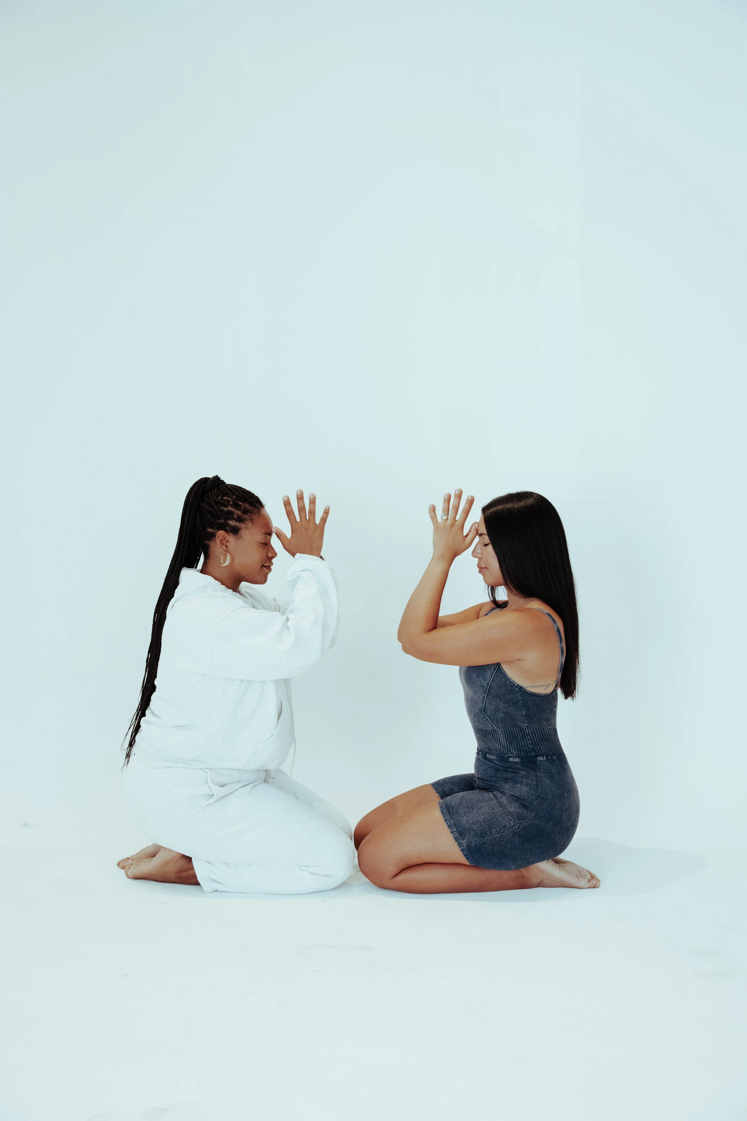 Two women kneeling and facing each other with their hands raised as if about to touch, on a plain white background.