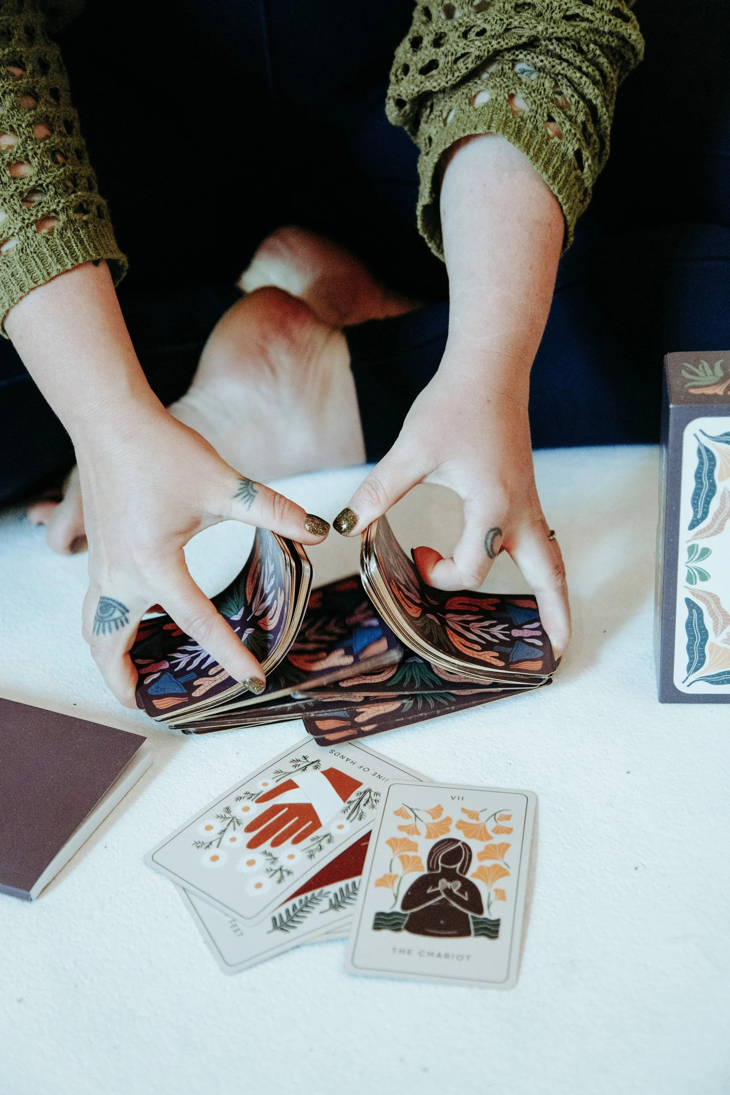 Person shuffling tarot cards on a white surface with tarot card boxes and drawn tarot cards visible, including 'The Chariot'.
