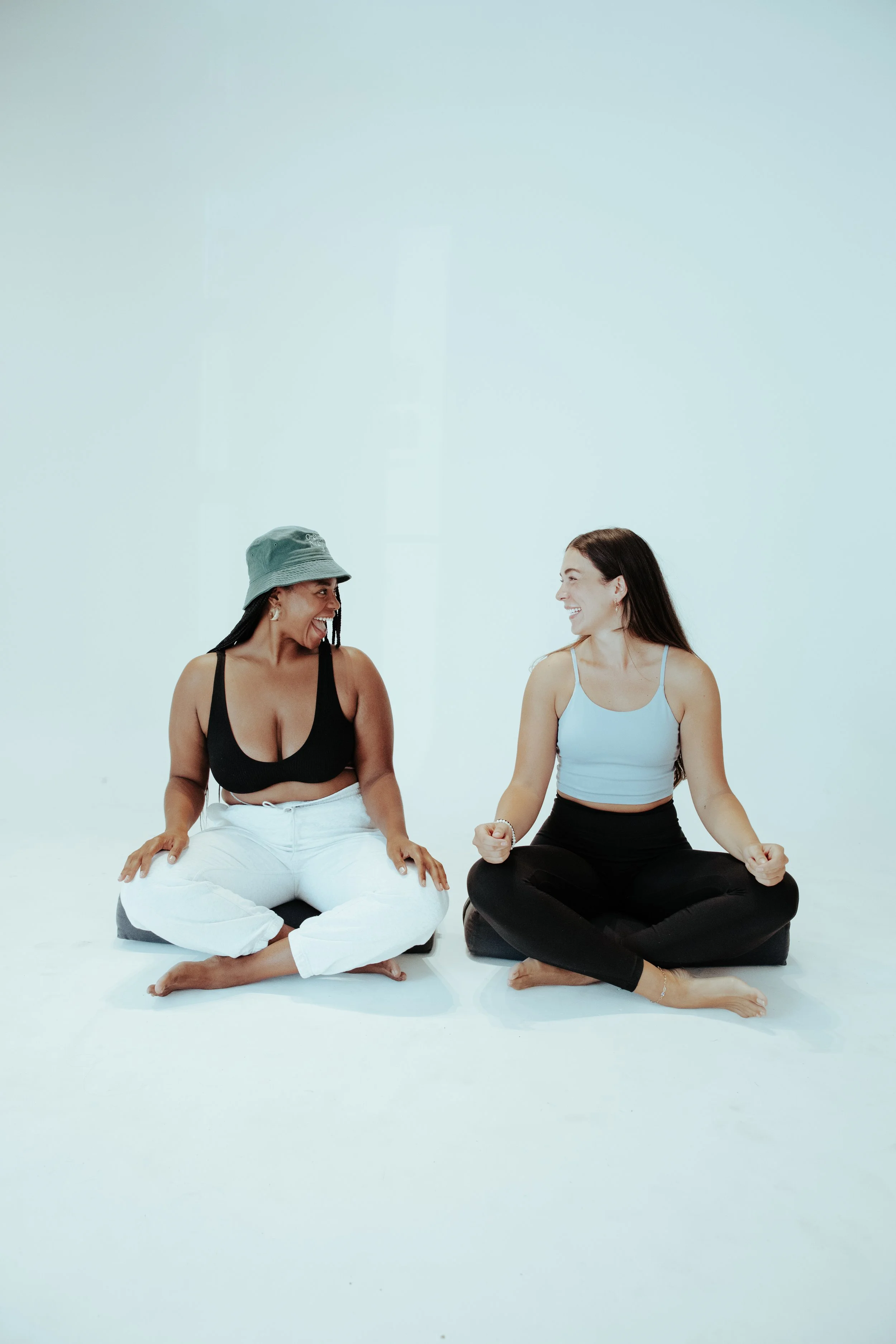 Two women sitting cross-legged on cushions, smiling and talking in a bright, minimal studio.