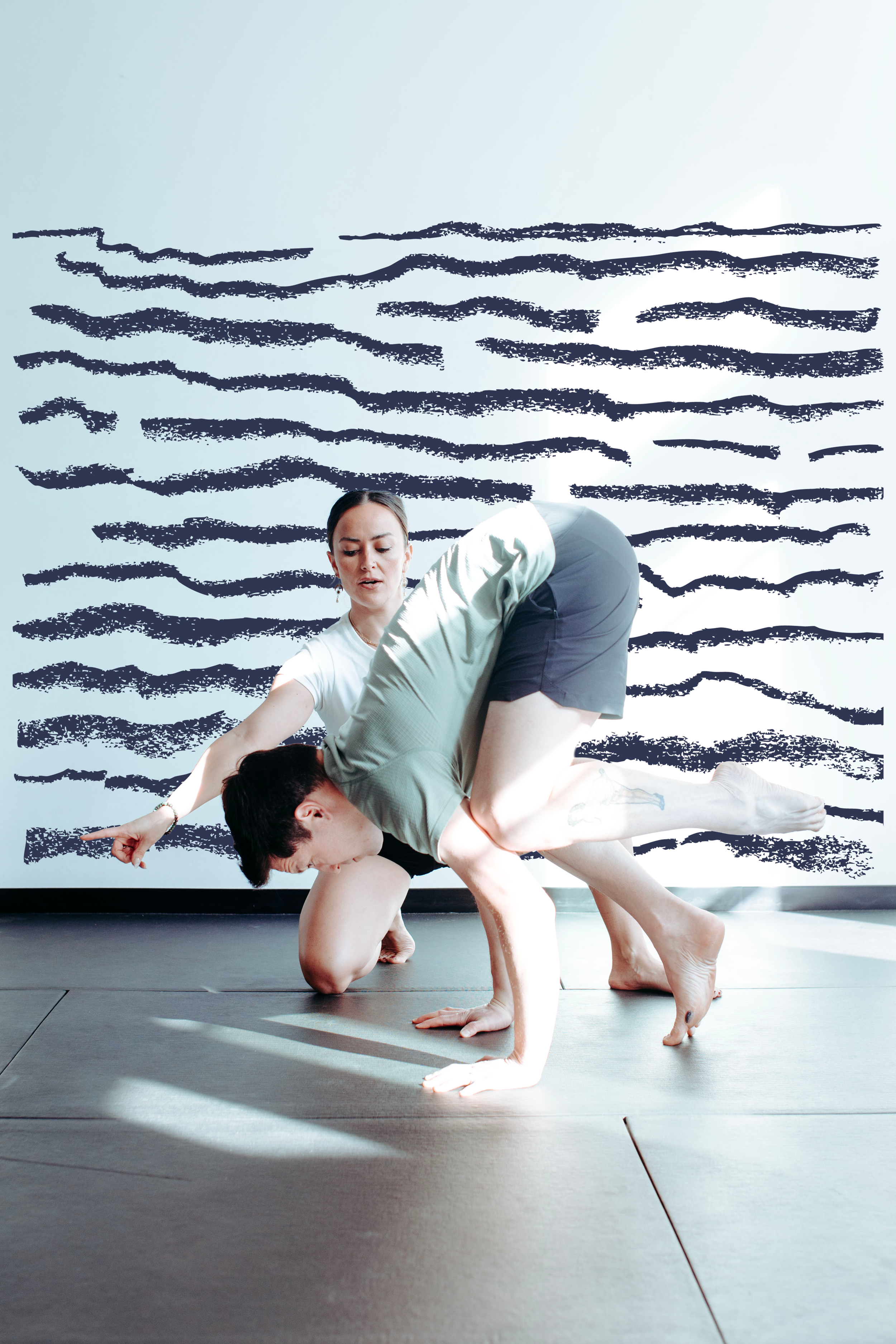 Two people practicing acro yoga indoors on a mat, with a gray wall featuring dark blue horizontal stripes in the background.