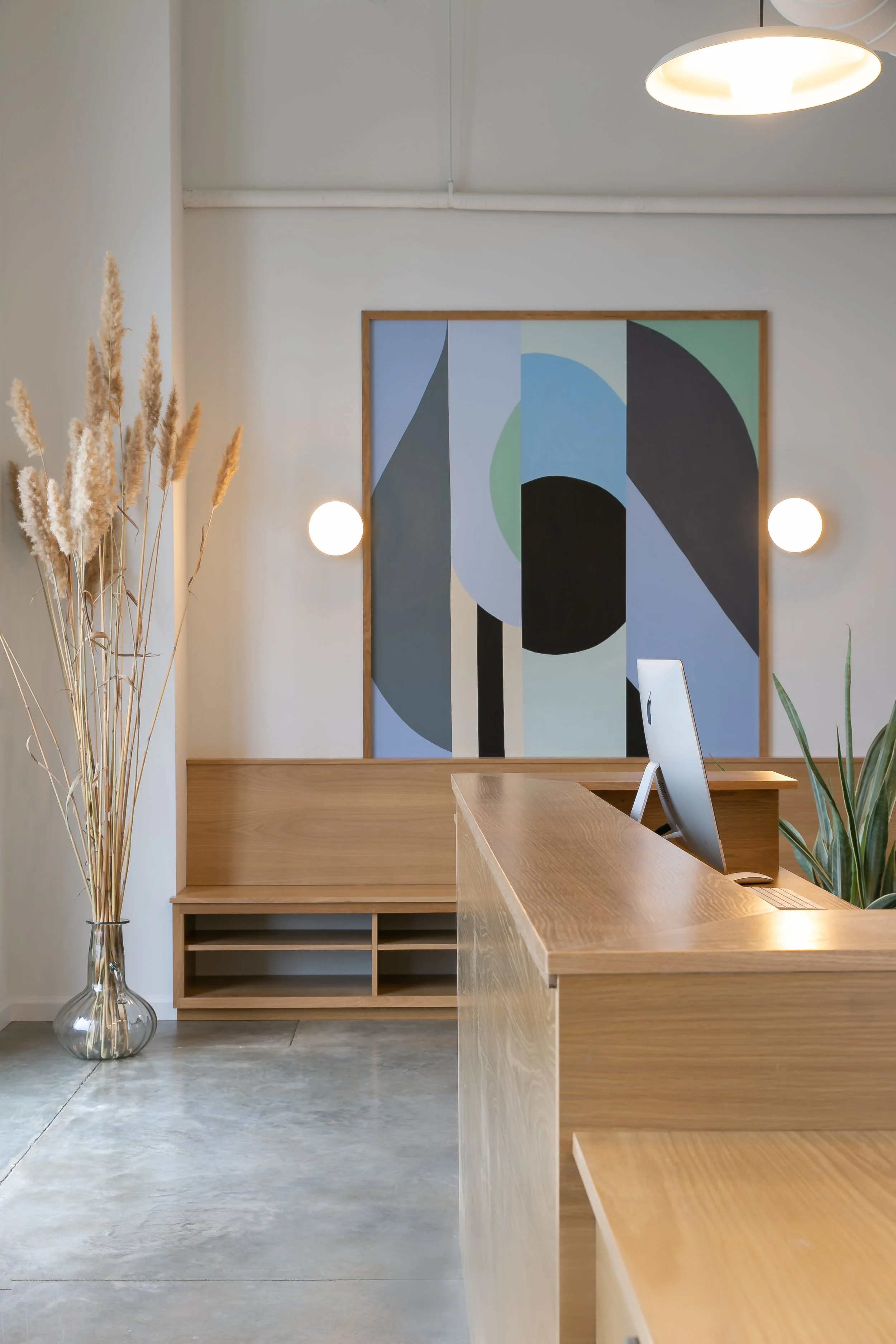Modern minimalist reception area with a wooden counter, an iMac computer, a large abstract geometric painting on the wall, soft globe wall lights, dried pampas grass in a glass vase, and houseplants.