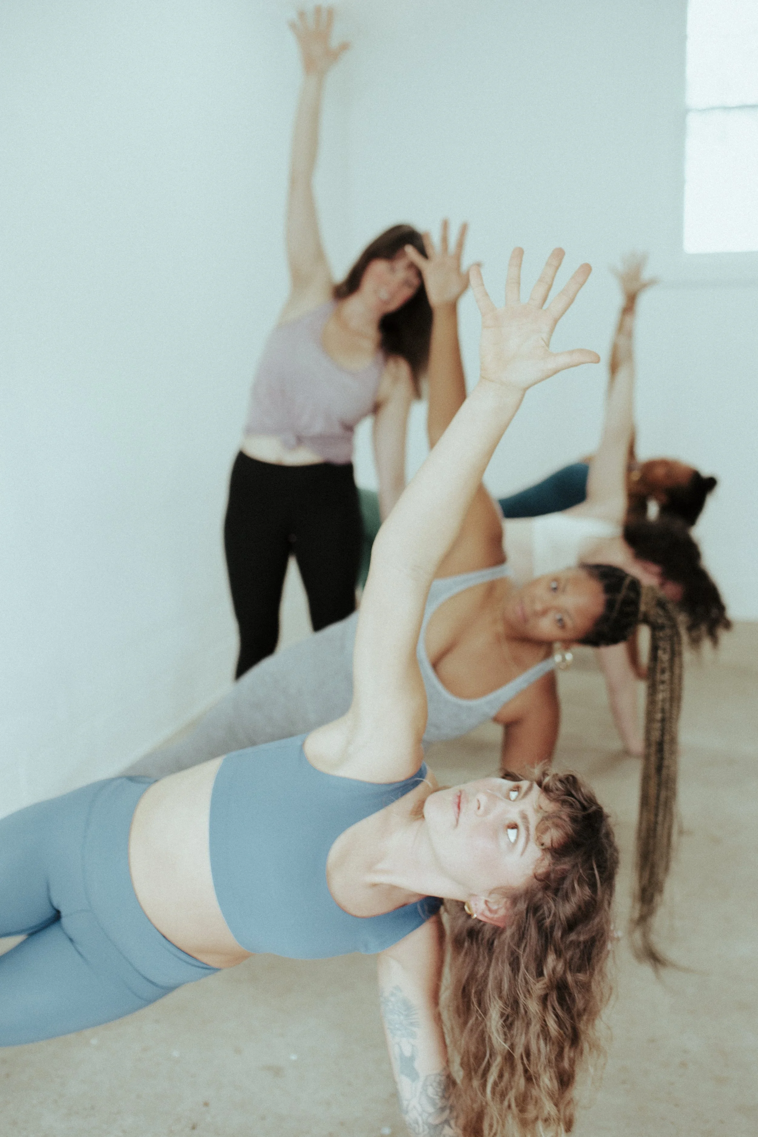 Group of women practicing yoga in a studio, with one woman in the foreground doing a side plank pose, and others in the background in various poses.