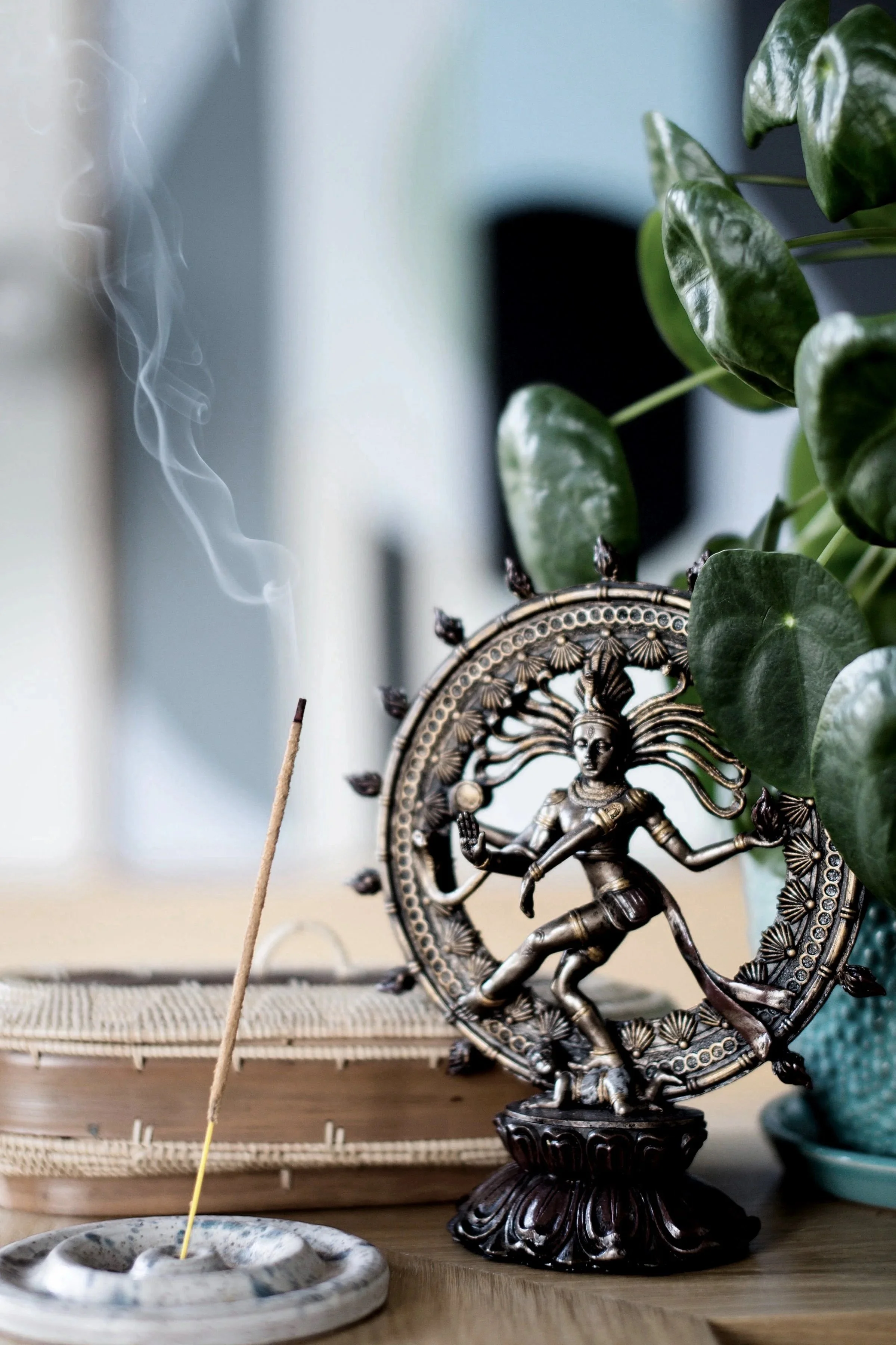 A metal statue of a dancing Hindu deity, possibly Lord Shiva, on a wooden surface next to a plant with green leaves. An incense stick is burning nearby, producing smoke.