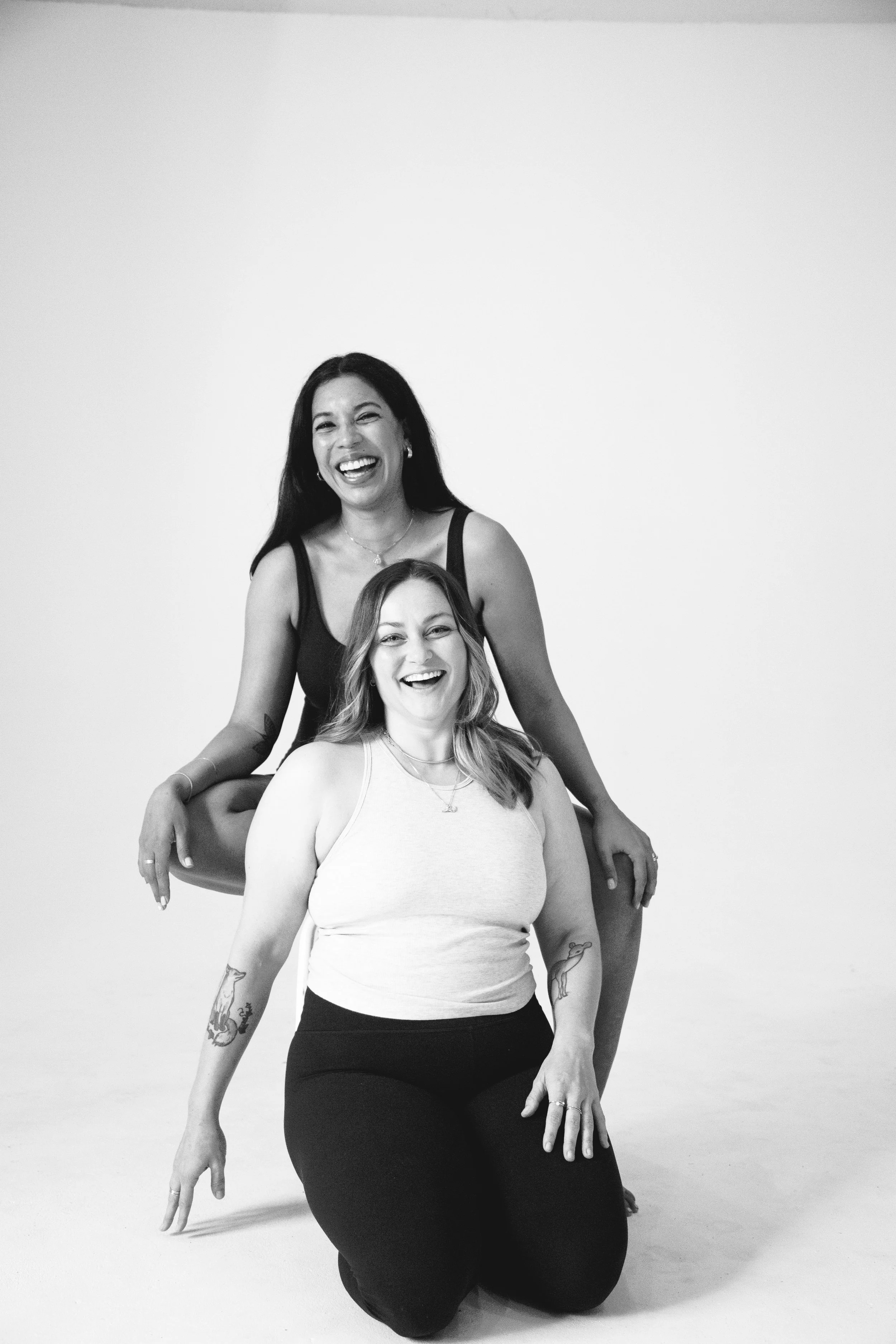 Two women smiling and laughing, one sitting on the floor and the other standing behind her, in a studio with a plain white background.