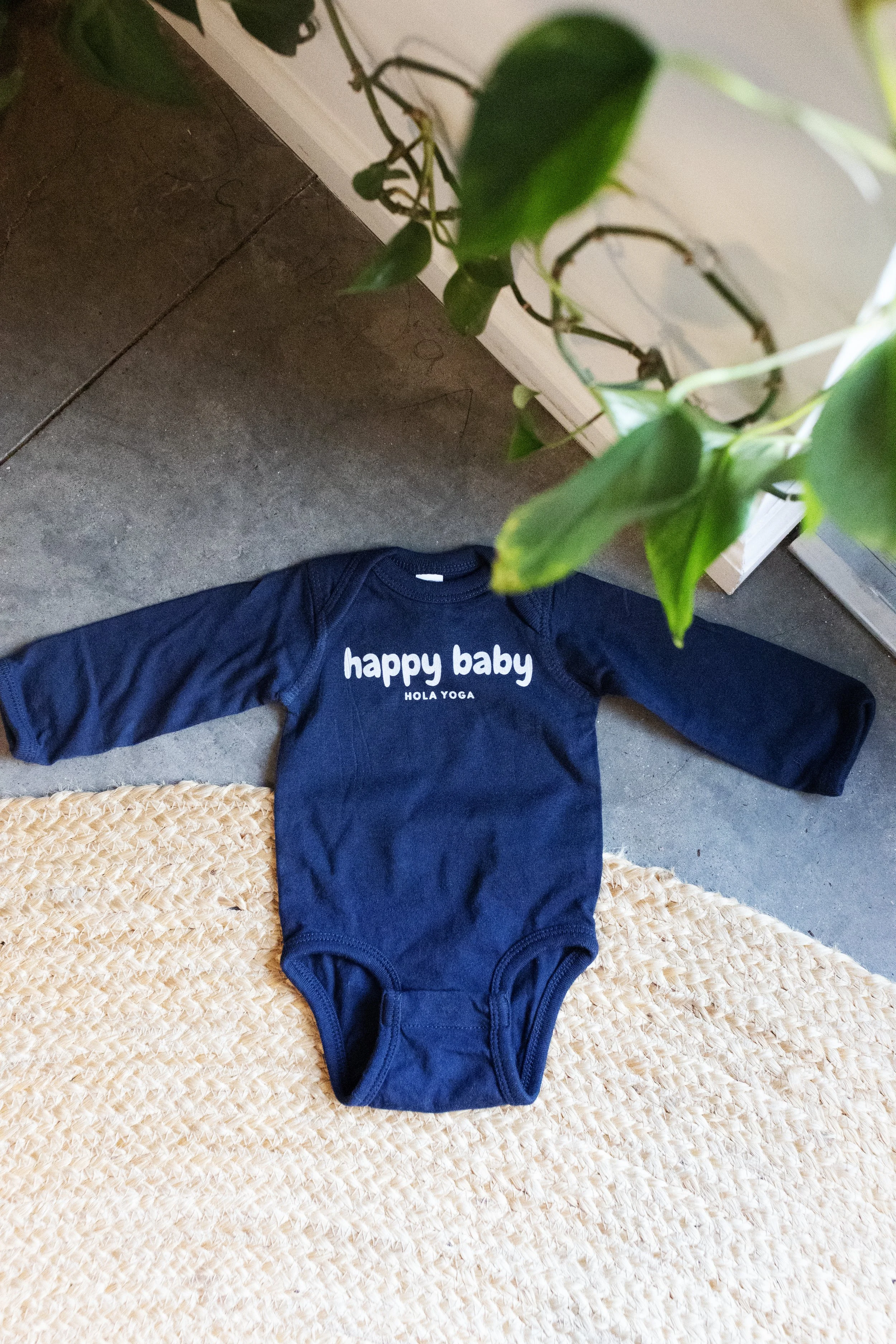 Blue baby bodysuit with white text that reads 'happy baby' and smaller text 'HOLA YOGA' underneath, lying on a beige woven rug on a concrete floor.