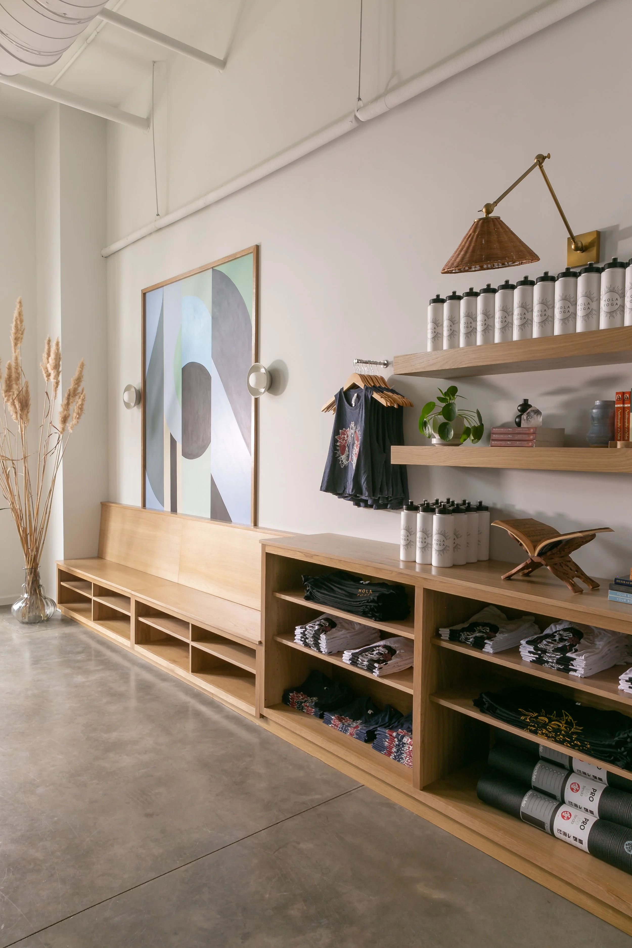 Interior of a fitness or yoga studio with wooden shelving and display of water bottles and athletic wear, modern artwork on the wall, and a tall vase with dried grass by the window.