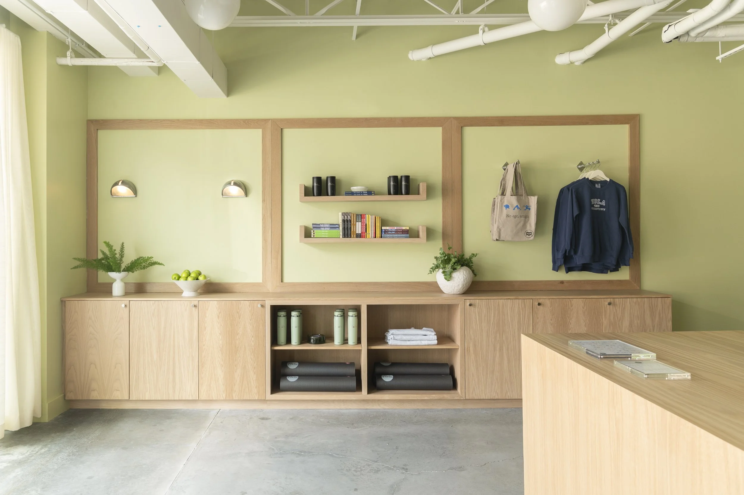 Modern reception area with a light wood desk, potted plants, white walls, and hanging ceiling lights, with the word 'HOLA' on the wall.