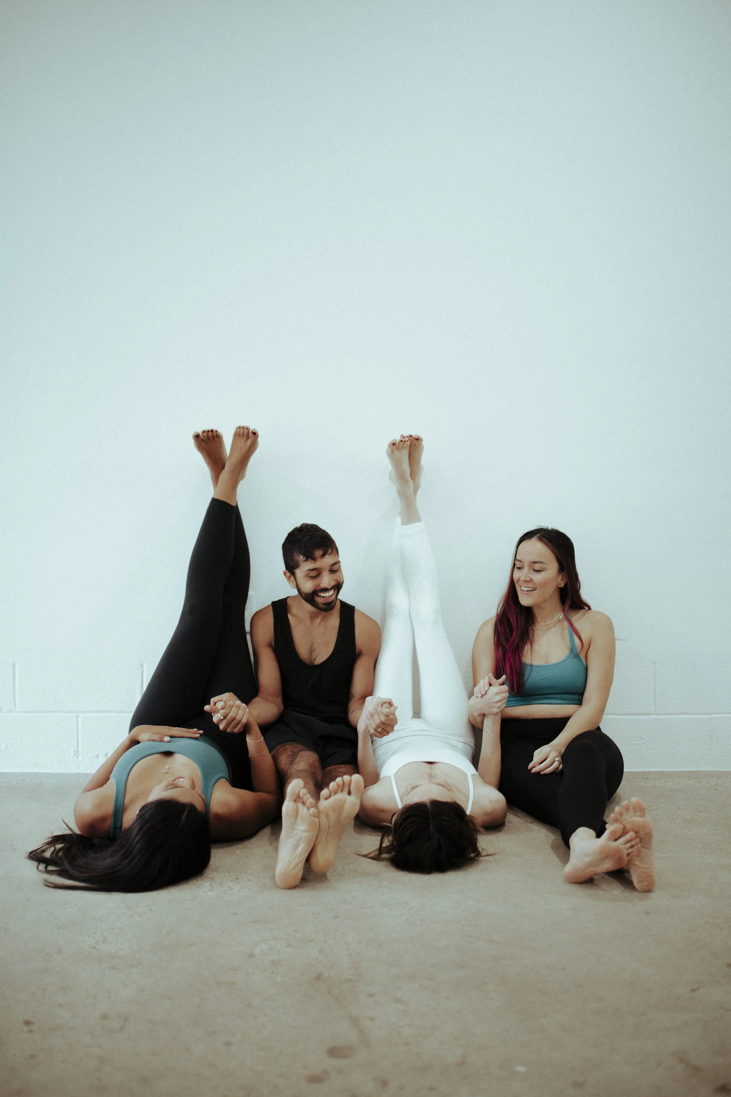 Four young adults sitting on the floor against a white wall, two women with their legs up against the wall and two men in the middle, holding hands and smiling.