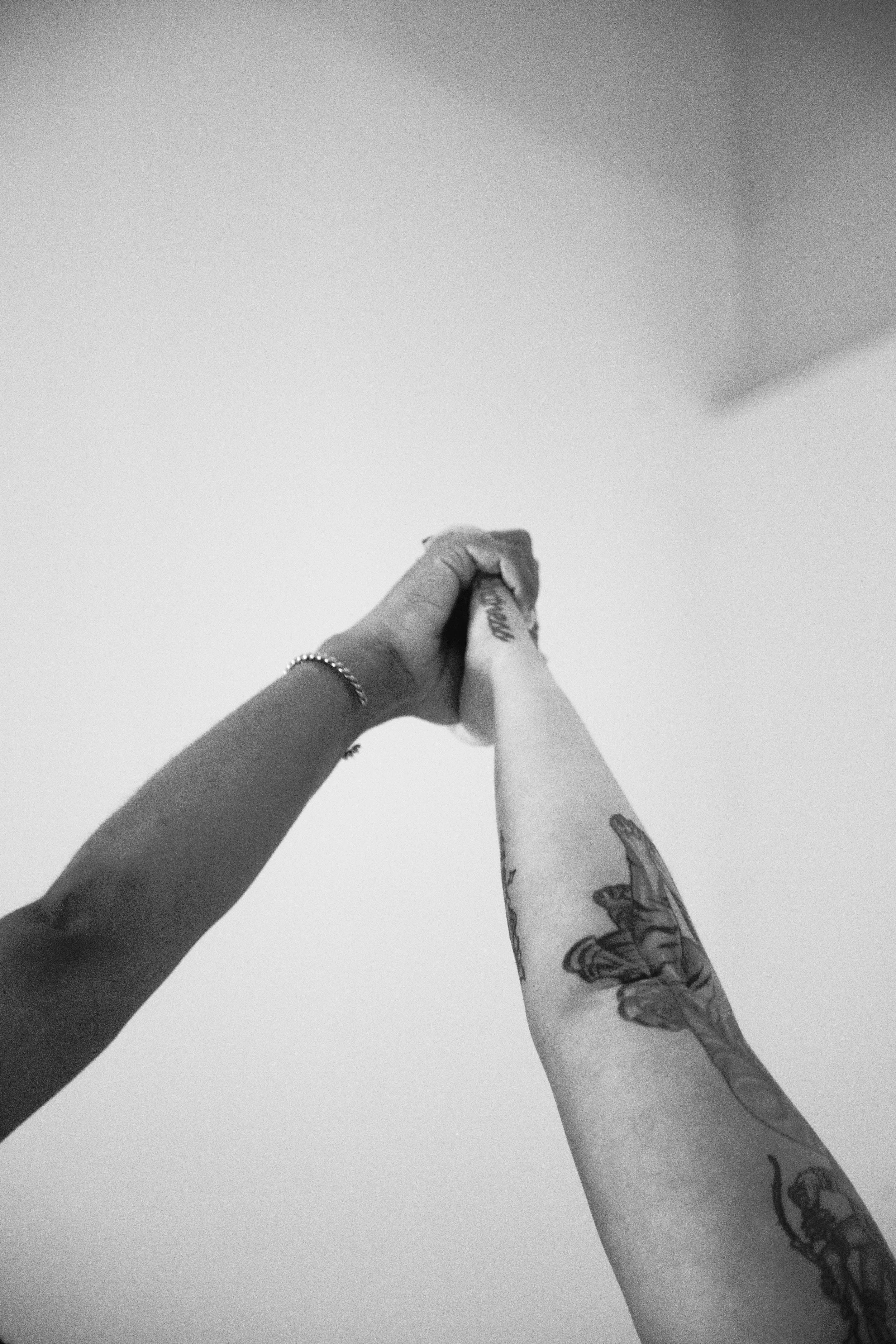 Two people holding hands, one with tattoos and a bracelet, against a plain background.