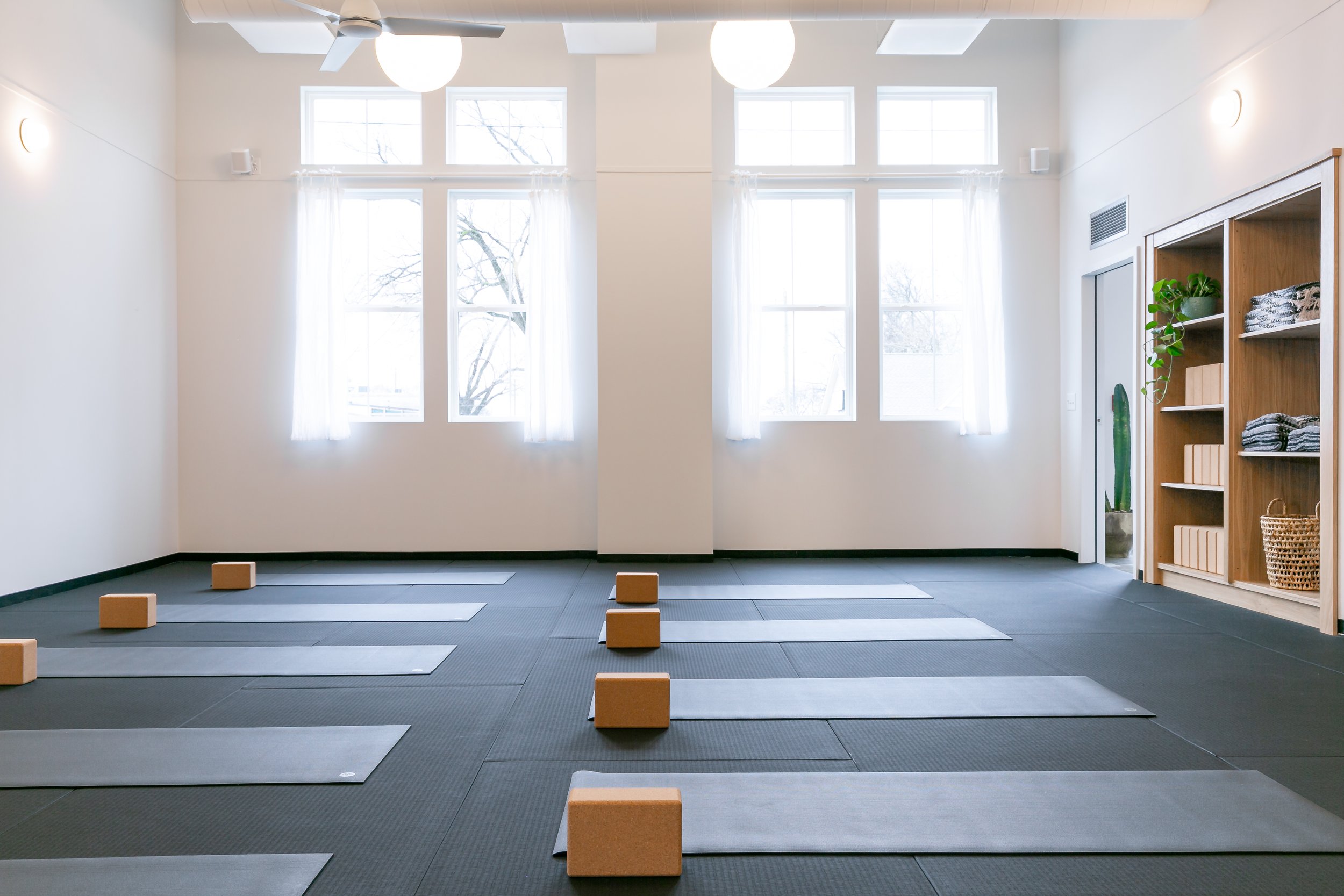 Yoga studio with mats and blocks, large windows with white curtains, and shelves with towels and plants.