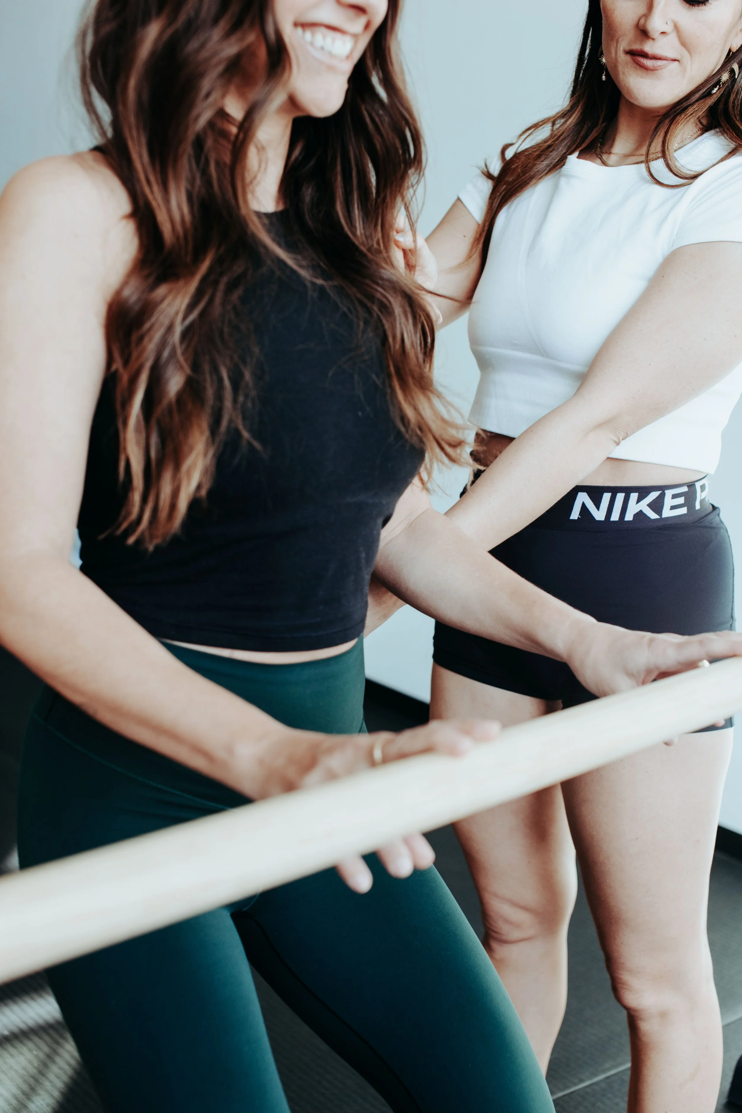 Two women standing near a ballet barre, engaged in a fitness or dance activity, wearing activewear, with smiles and a relaxed atmosphere.