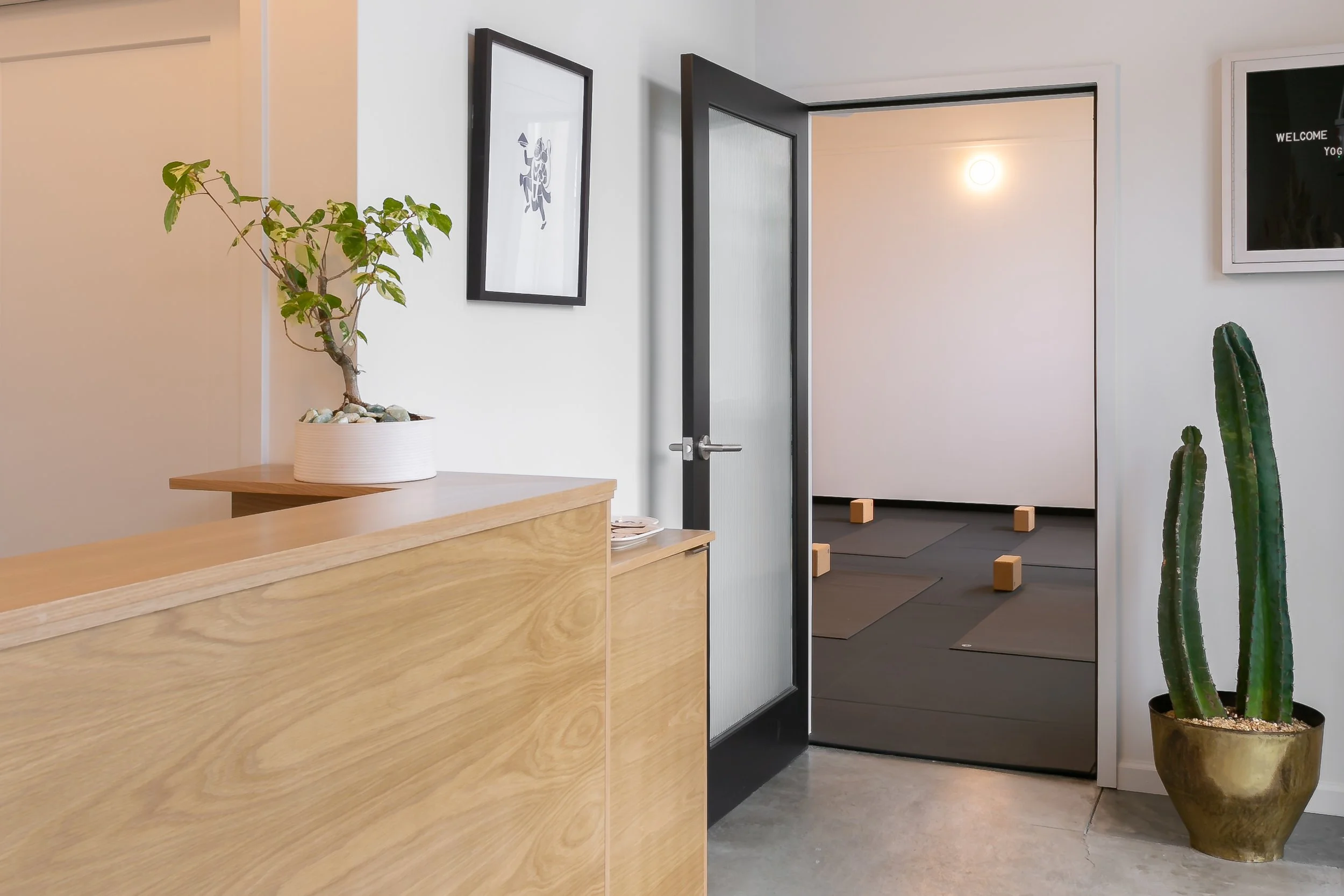 Interior of a wellness or yoga studio with a reception desk, a plant, wall art, and a room with yoga mats and wooden blocks.