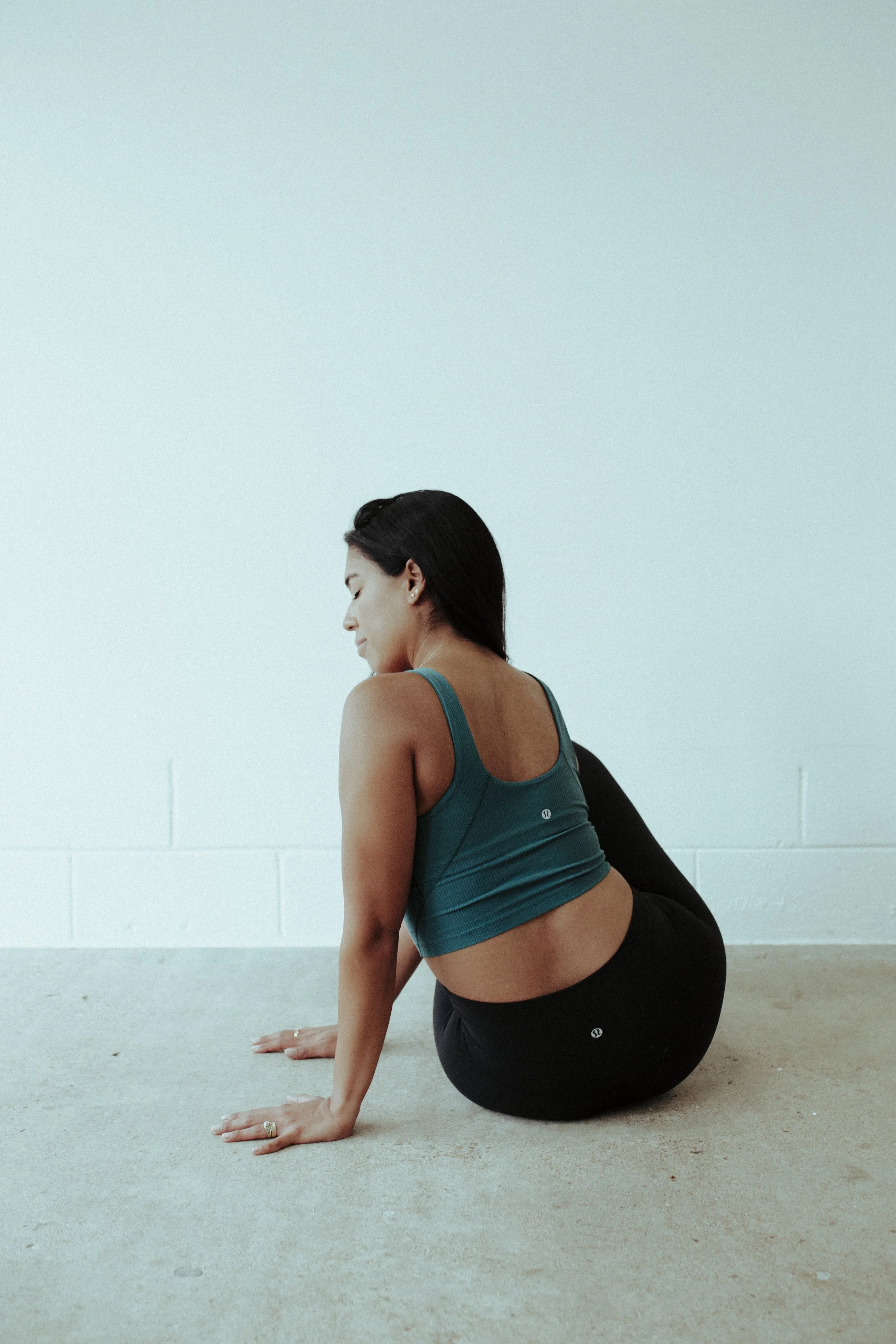 A woman practicing yoga on the floor against a white wall, sitting with her legs bent and arms supporting her body, wearing a teal athletic top and black leggings.