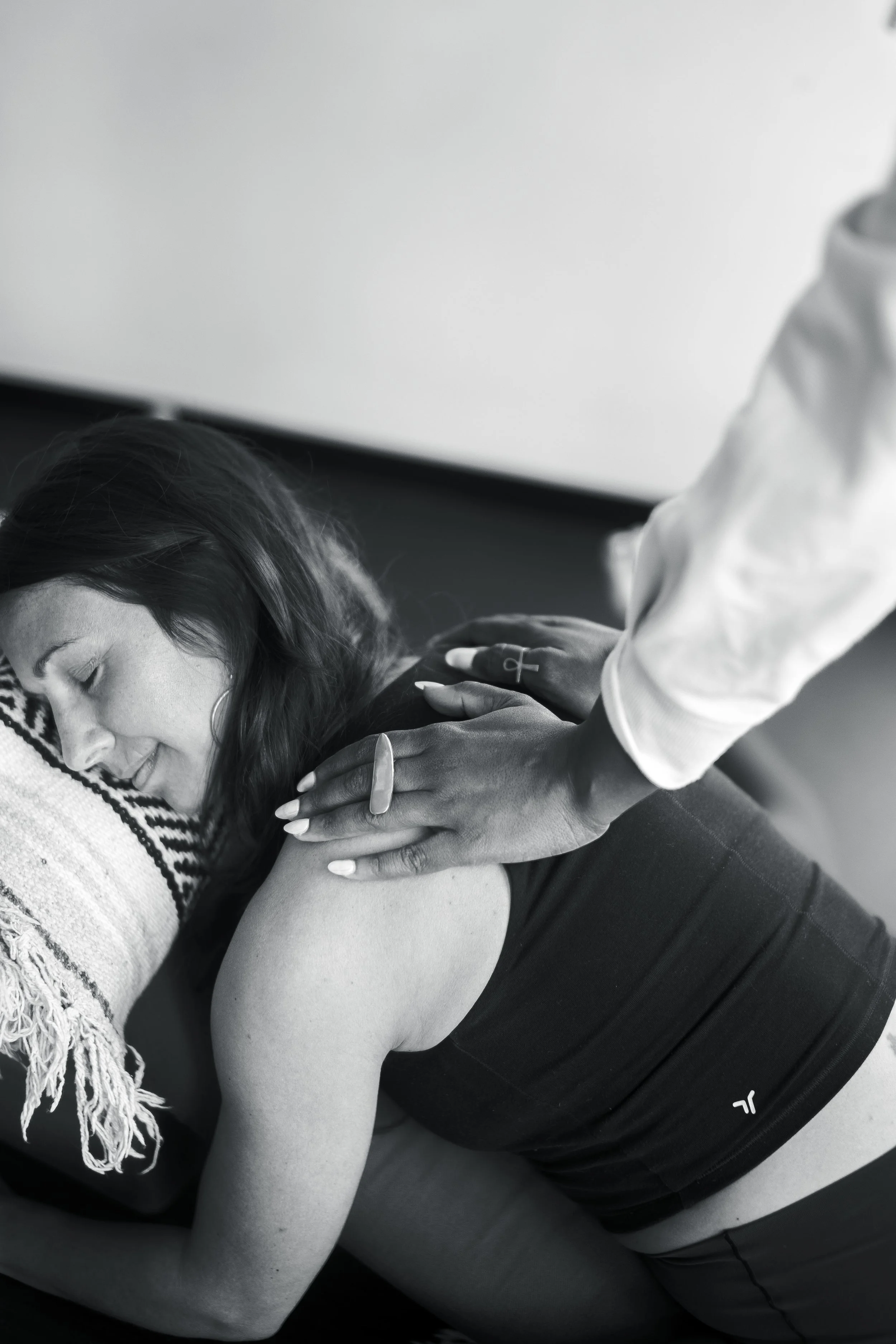 A woman is lying face down on a massage table, receiving a back massage from a therapist whose hands are on her shoulder and upper back, in a peaceful setting.
