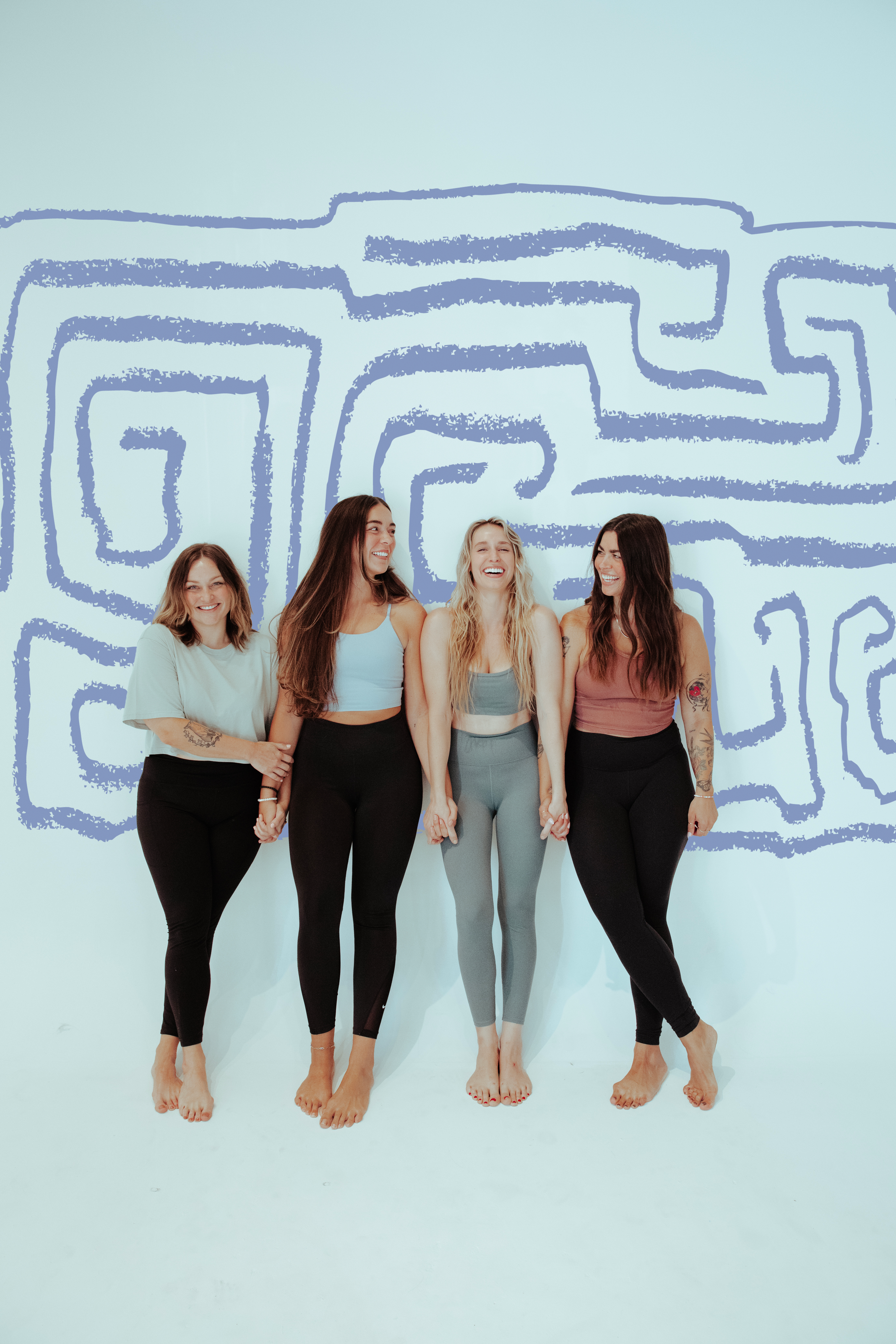 Four women holding hands and smiling in front of a blue maze wall art, dressed in athletic wear.