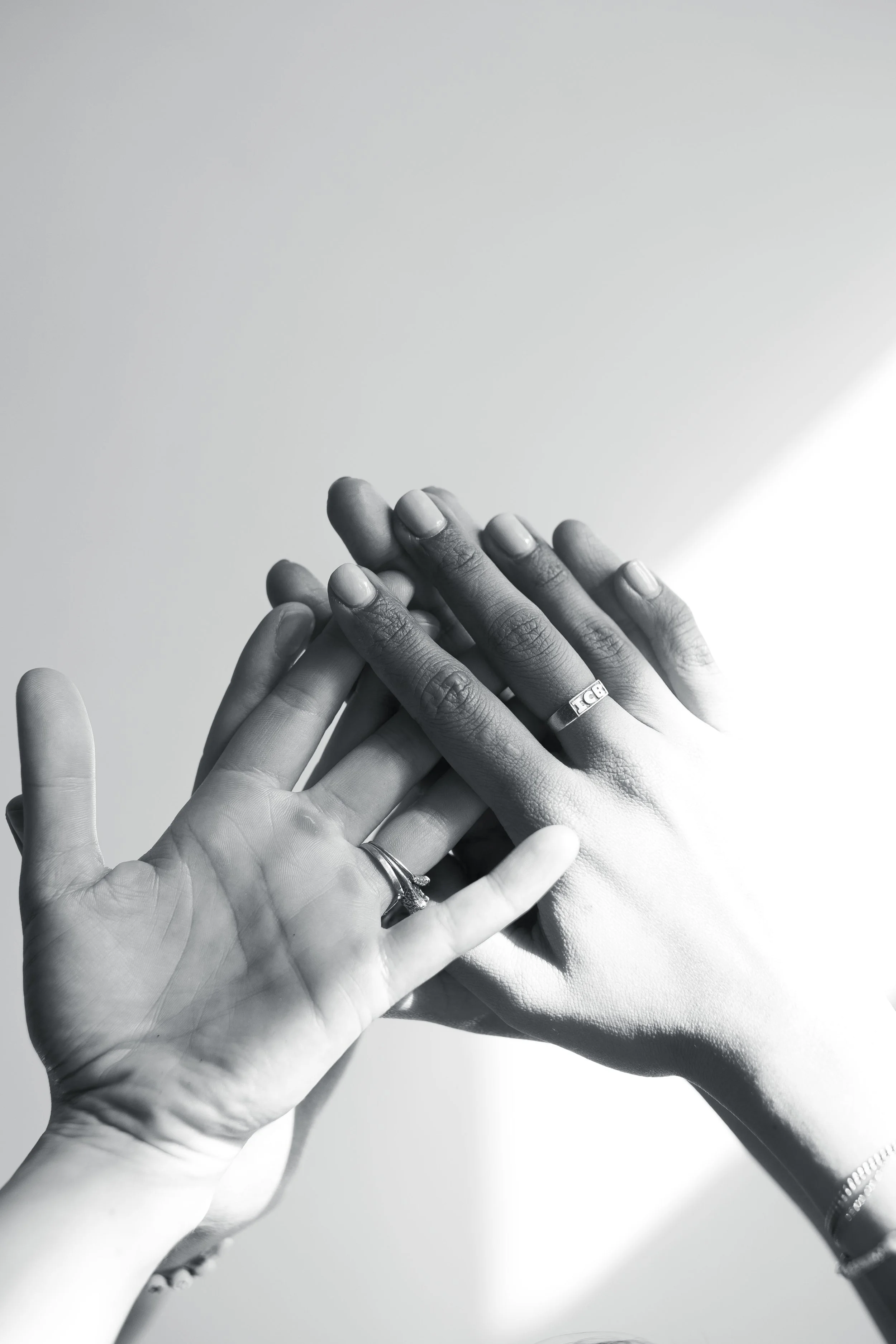 Black and white photograph of multiple hands stacked together, each with a ring, against a plain background.