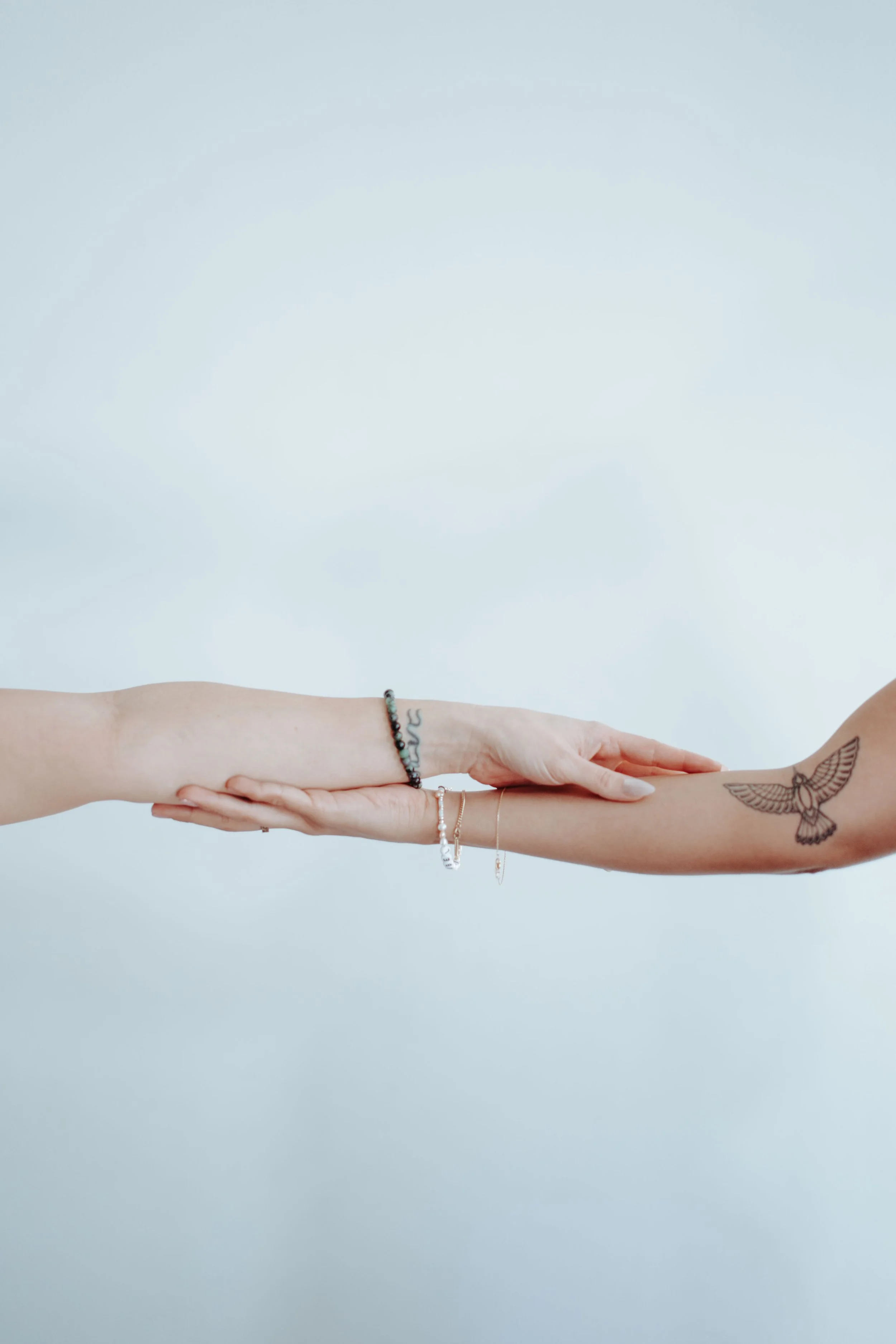 Two hands touching, one with a tattoo of a bird on the forearm, the other with jewelry including a bracelet and rings, against a plain light background.