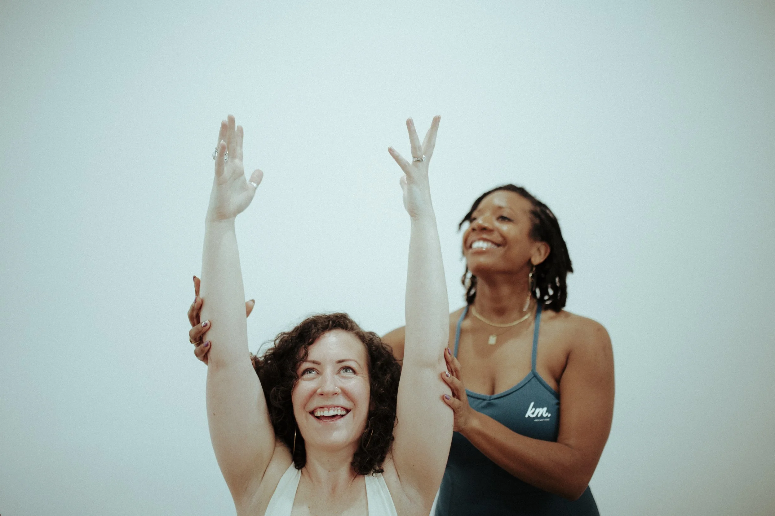 A woman practicing yoga, raising her arms in a yoga pose while an instructor assists her from behind, in a light-colored room.
