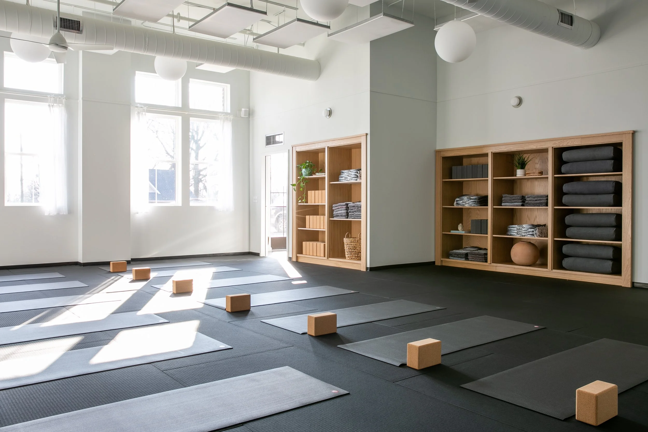 Yoga studio with mats, blocks, and shelves of blankets and towels, illuminated by natural light from large windows.