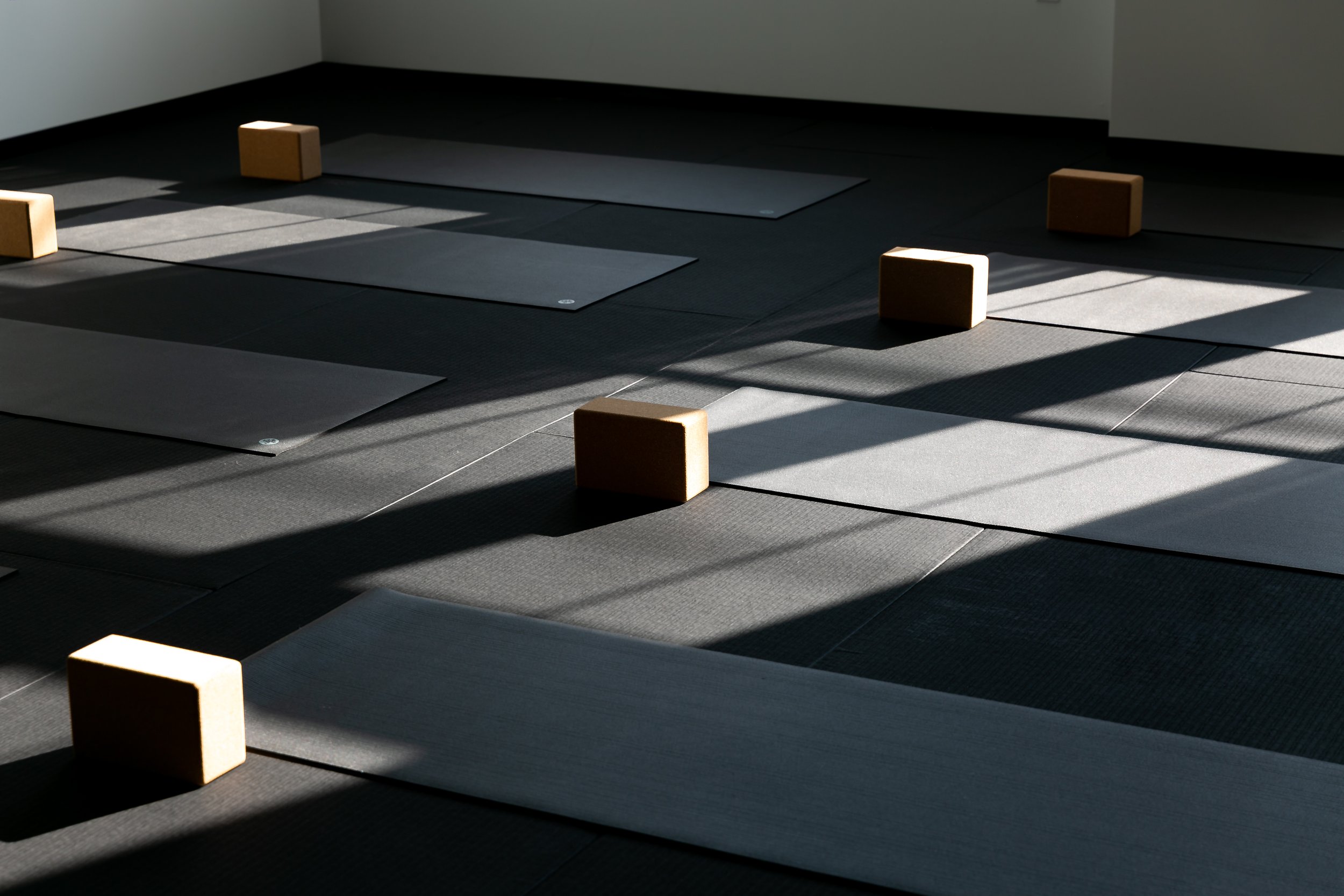 Yoga mats and wooden blocks arranged on a black floor with sunlight streaming in, creating shadows.