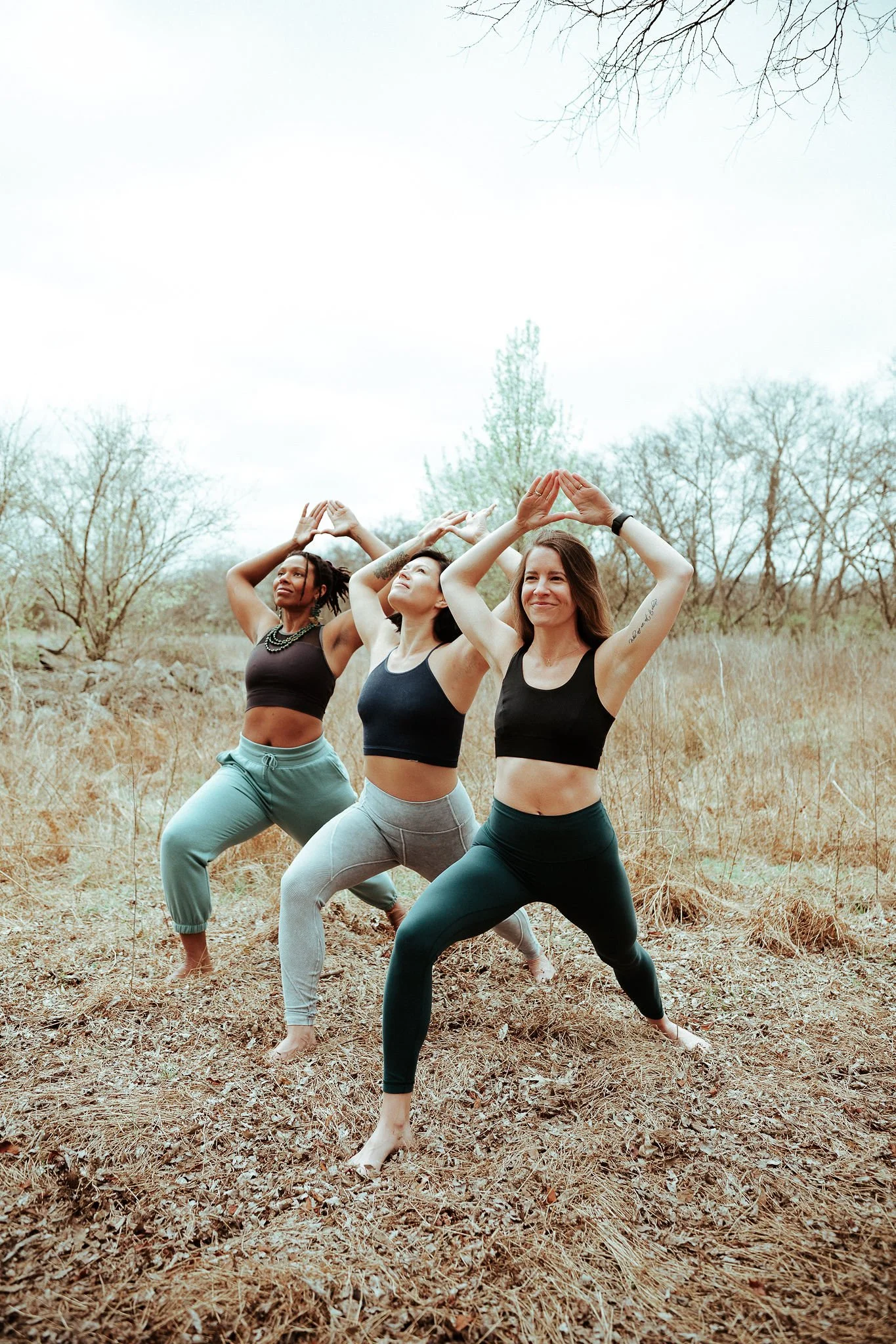 Three women practicing yoga outdoors on a leaf-covered field with bare trees in the background, standing in wide-legged stance with arms raised overhead in a prayer position.